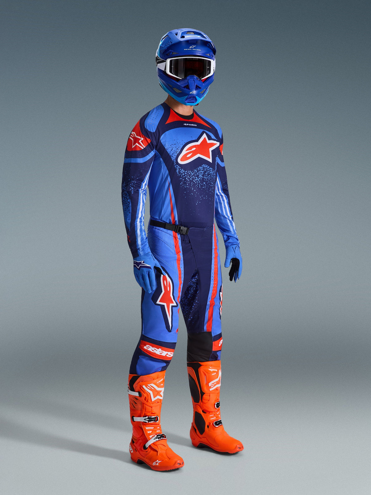 Zawodnik motocrossowy w kompletnym stroju wyścigowym, niebiesko-granatowa koszulka Techstar Nomur z pomarańczowym logo Alpinestars, dopasowane niebiesko-granatowe spodnie z pomarańczowymi akcentami, niebieski kask z białymi goglami, jaskrawopomarańczowe buty i niebieskie rękawice, skoordynowany zestaw do wyścigów terenowych