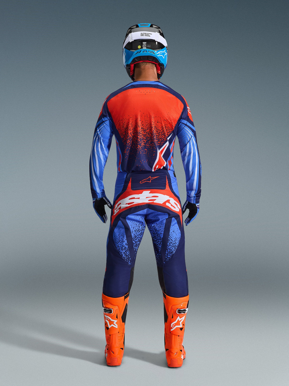 Kierowca motocrossowy widziany od tyłu w koszulce Techstar Nomur 2026 i pasujących spodniach, kolorystyka Dark Navy i Orange z akcentami UCLA Blue, jaskrawopomarańczowe buty, niebiesko-biały kask i czarne rękawice, kompletny profesjonalny strój do wyścigów off-road