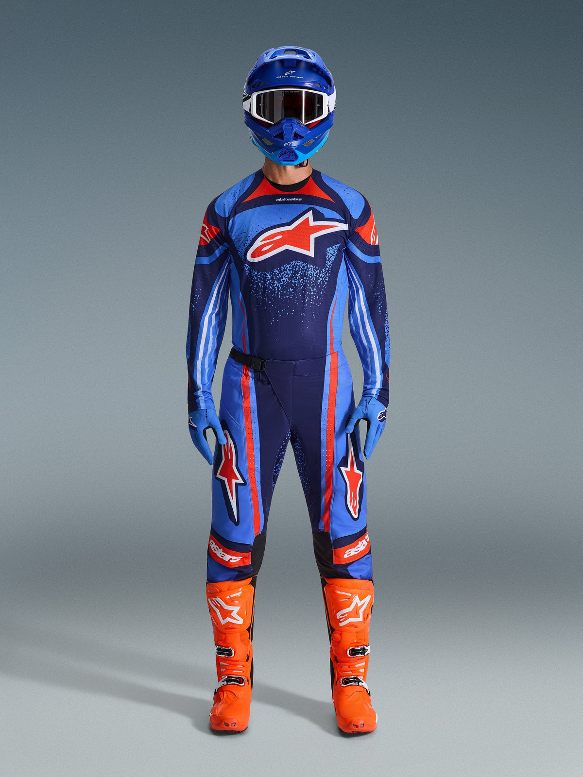 Zawodnik motocrossowy ubrany w koszulkę Alpinestars 2026 Techstar Nomur w kolorach granatowym, pomarańczowym i błękitnym, w zestawie z dopasowanymi spodniami, niebieskim kaskiem z goglami oraz jaskrawopomarańczowymi butami wyścigowymi, kompletny profesjonalny strój off-road