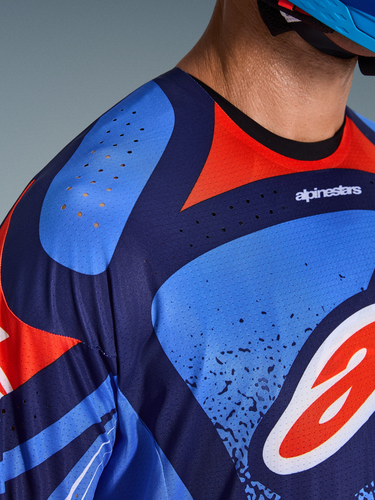 Zbliżenie na koszulkę Alpinestars 2026 Techstar Nomur, koszulka motocrossowa, ciemnogranatowa, pomarańczowa i błękitna (UCLA Blue), z laserowo wyciętymi otworami wentylacyjnymi na ramieniu i klatce piersiowej, techniczny materiał odprowadzający wilgoć, sportowy krój wyścigowy