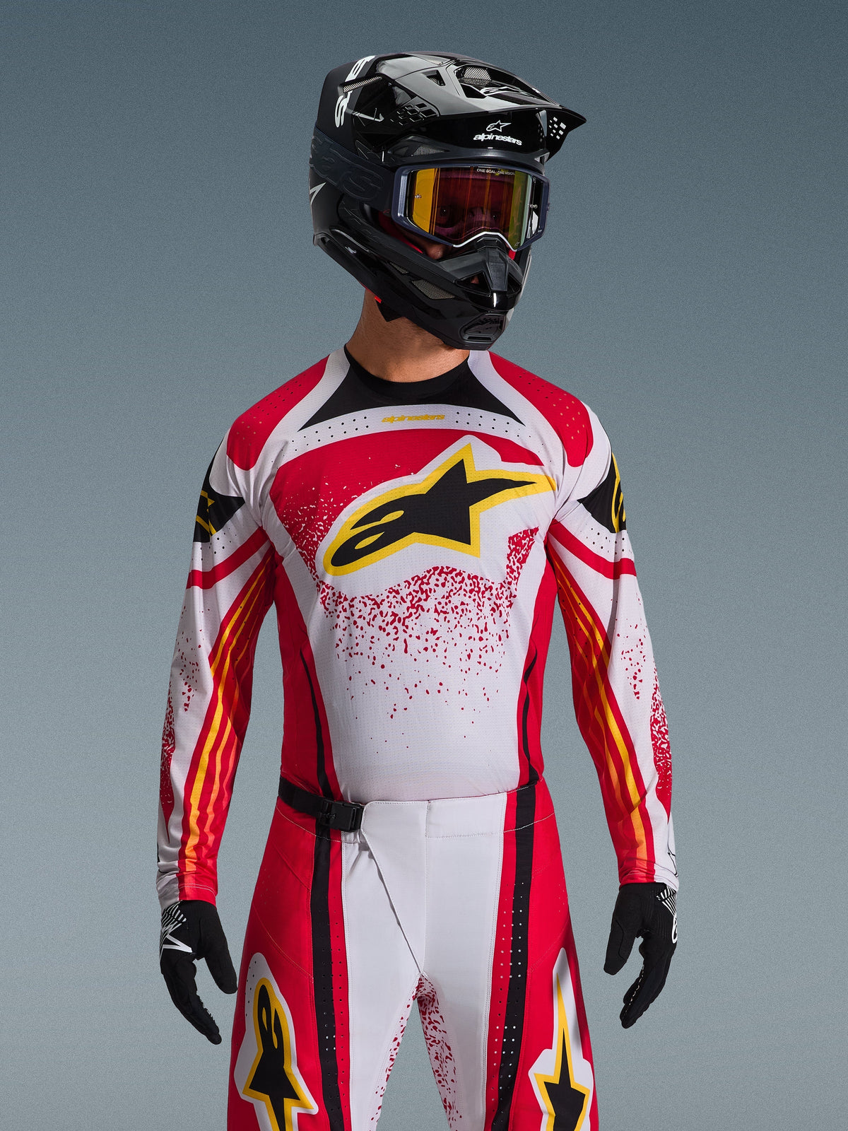 Alpinestars 2026 Techstar Nomur Jersey, koszulka motocrossowa, czerwona, biała i czarna, z dużym czarnym logo A-star na piersi z żółtymi akcentami, czerwony wzór typu splatter, noszona z czarnym kaskiem i dopasowaną odzieżą off-road
