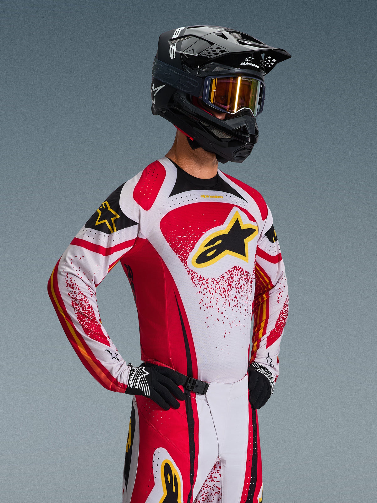 Zawodnik motocrossowy w koszulce Alpinestars 2026 Techstar Nomur, czerwono-biało-czarnej z żółtymi akcentami, w zestawie z dopasowanymi spodniami, czarnym kaskiem ze złotymi goglami i czarnymi rękawicami, atletyczny krój wyścigowy do profesjonalnej rywalizacji off-road