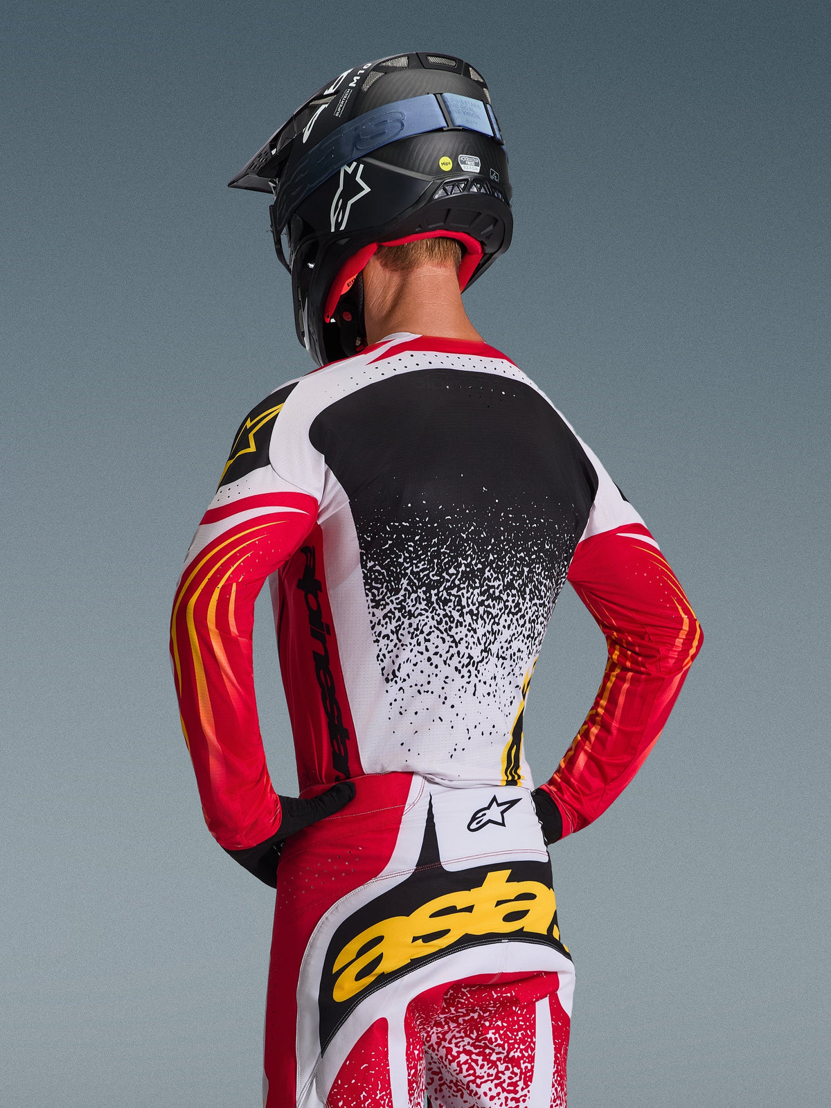Motocyklista motocrossowy widziany od tyłu w koszulce Alpinestars 2026 Techstar Nomur i dopasowanych spodniach, czerwono-biało-czarny wzór z żółtymi akcentami i nakrapianym gradientem na plecach, czarny kask Alpinestars i czarne rękawice, profesjonalny zestaw odzieży do wyścigów off-road
