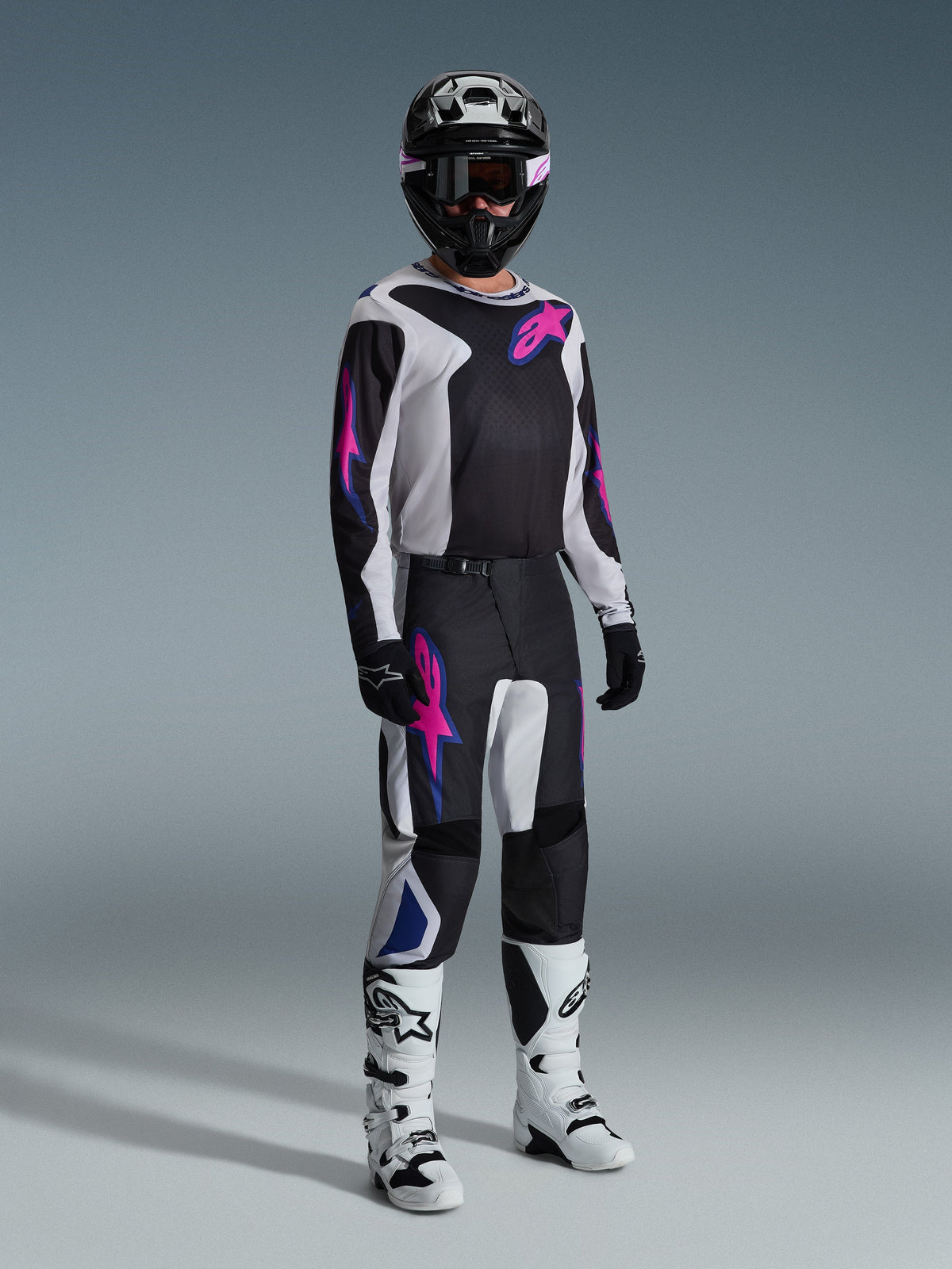 Motocross rider wearing complete racing gear, 2026 Fluid Grid Jersey in black with light gray and purple accents, matching Fluid Grid Pants, black helmet with coordinated goggles, white and black boots, and black gloves, professional studio setup for off-road racing.