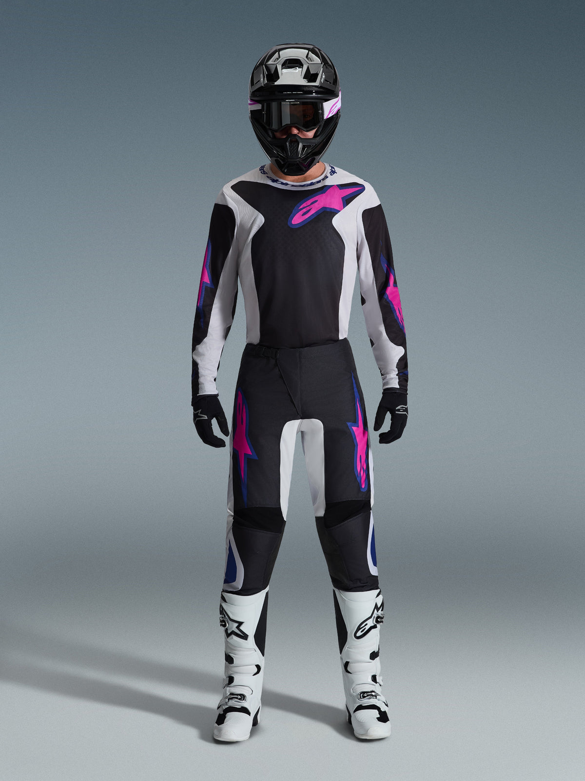 Motocross rider wearing complete racing gear, Alpinestars 2026 Fluid Grid Jersey and matching pants in black and light gray with vibrant purple accents, black helmet with purple goggles, white motocross boots, and black gloves, coordinated outfit for off-road performance
