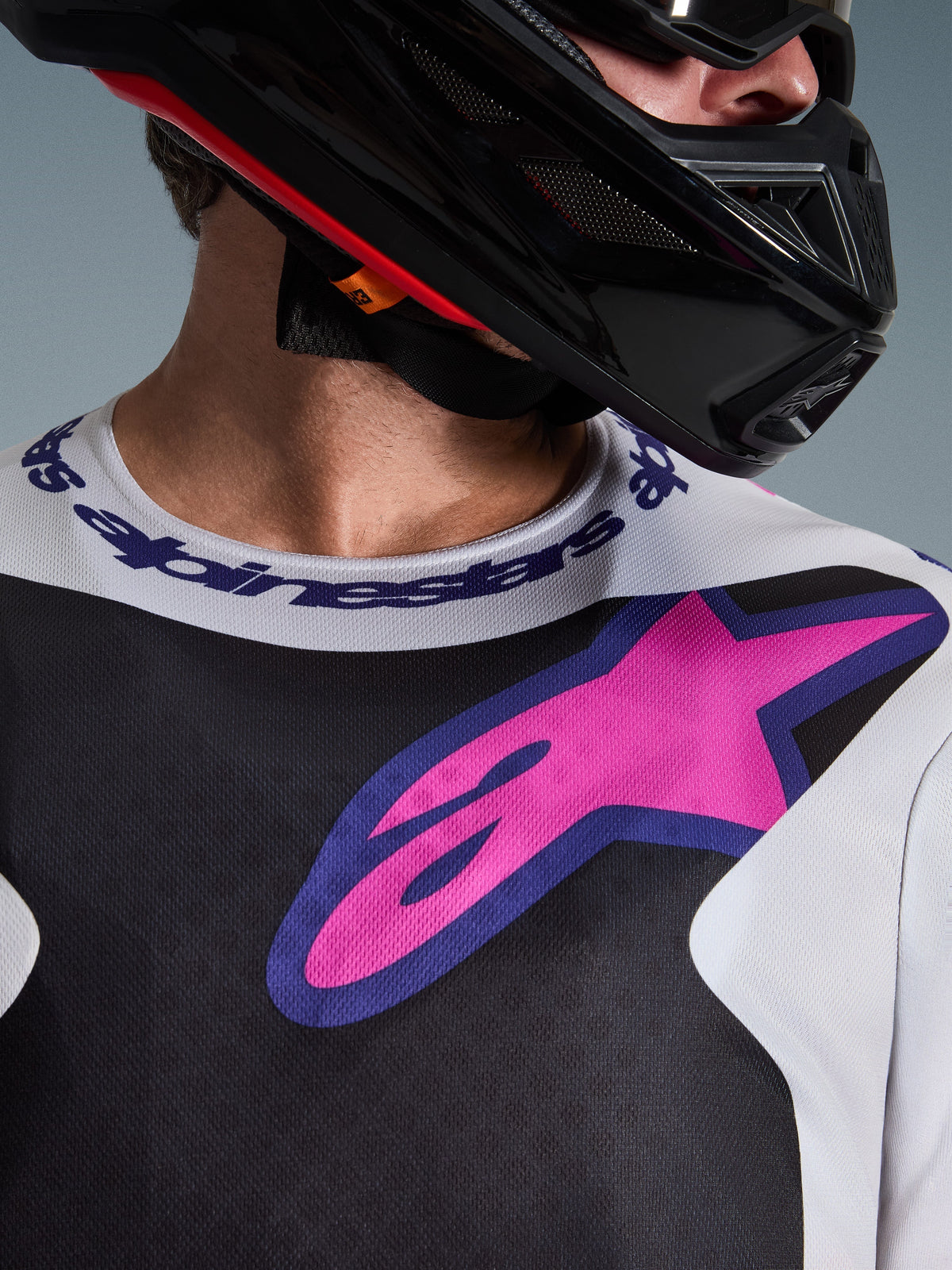 Alpinestars 2026 Fluid Grid Jersey, Motocross Jersey, Black Light Gray and Purple, close-up of chest and neck area featuring large pink star logo, branded sport collar, worn with black helmet, performance off-road gear