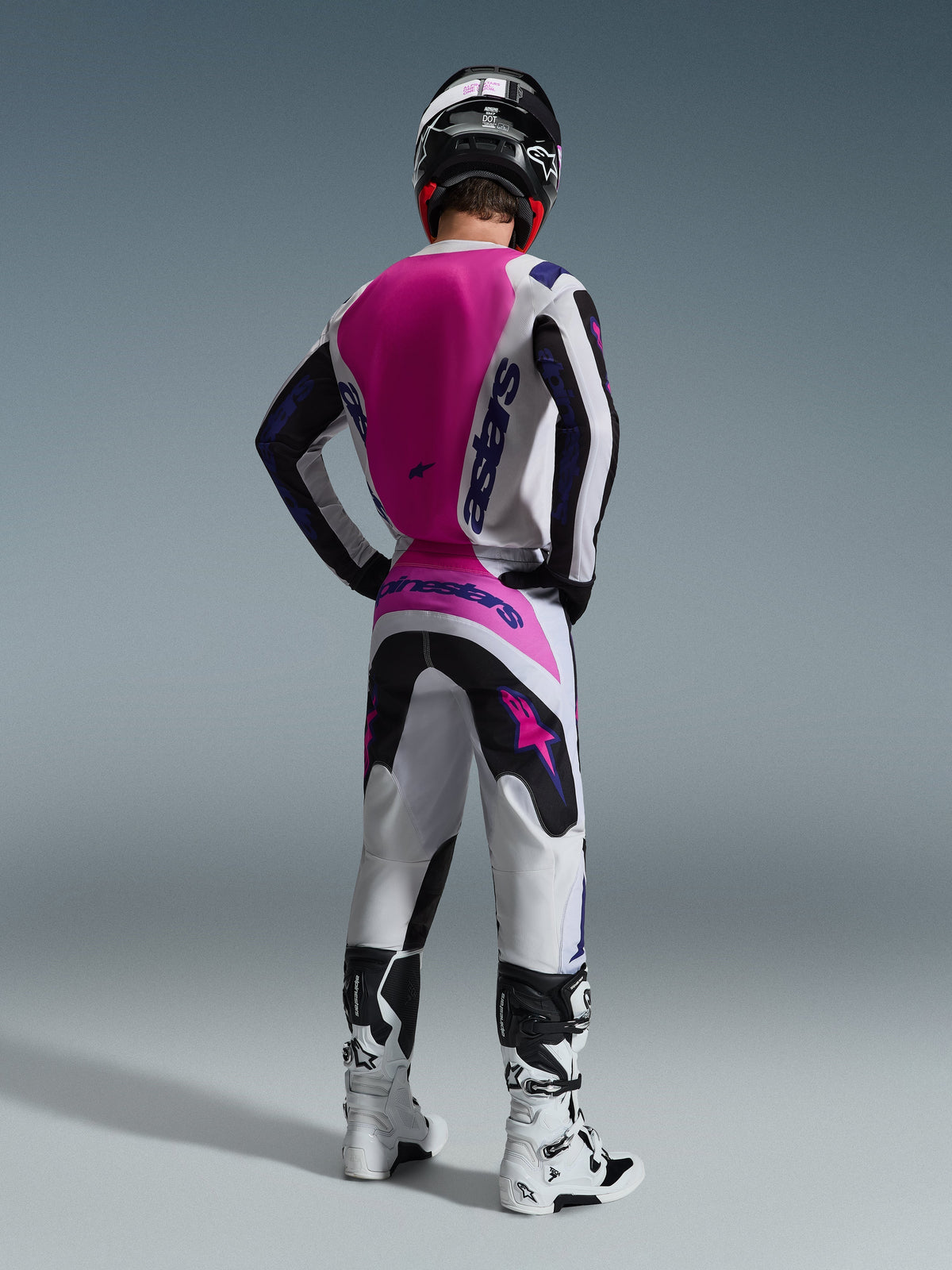 Alpinestars 2026 Fluid Grid Gear Set, zawodnik motocrossowy widziany od tyłu, biała bluza z różowym panelem na plecach i fioletowym logo, dopasowane biało-czarne spodnie z różowymi akcentami, biało-czarne buty i czarny kask, skoordynowany strój do wyścigów terenowych