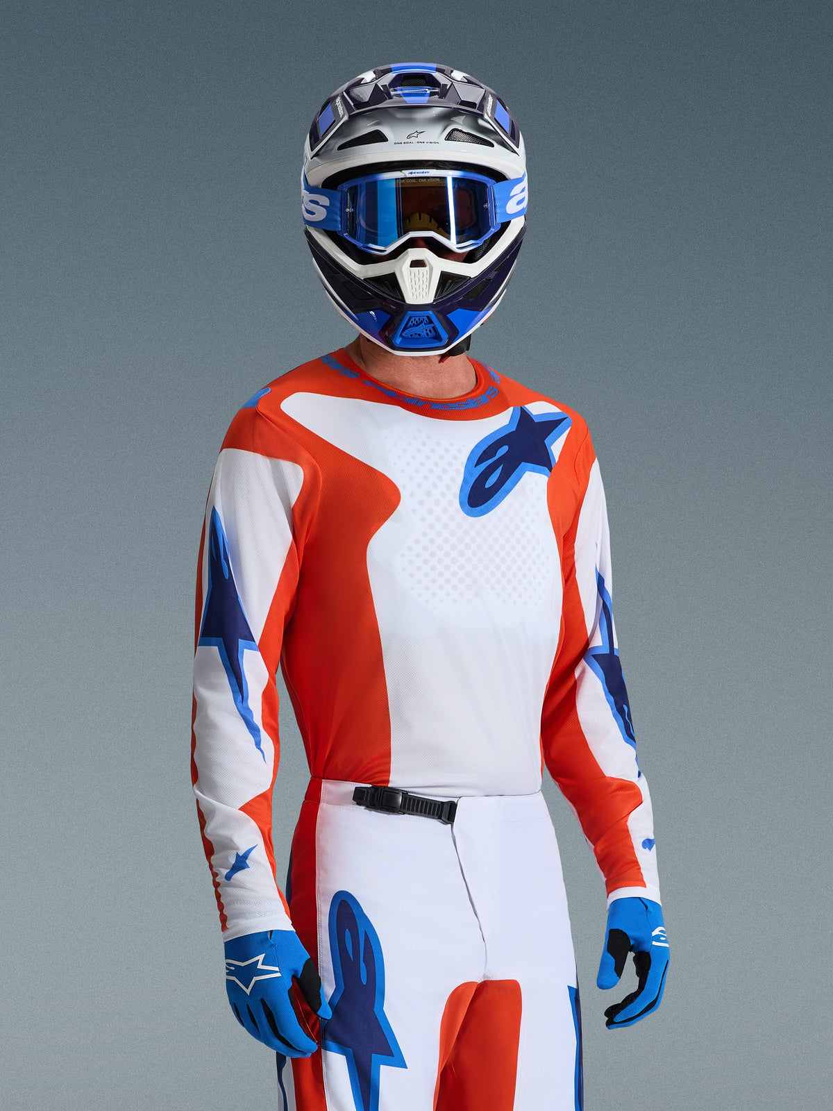 Koszulka Alpinestars Fluid Grid 2026, koszulka motocrossowa w kolorze Orange UCLA Blue, noszona przez motocyklistę z dopasowanymi biało-pomarańczowymi spodniami, niebiesko-białym kaskiem, niebieskimi goglami i niebieskimi rękawicami, kompletny zestaw wyścigowy off-road
