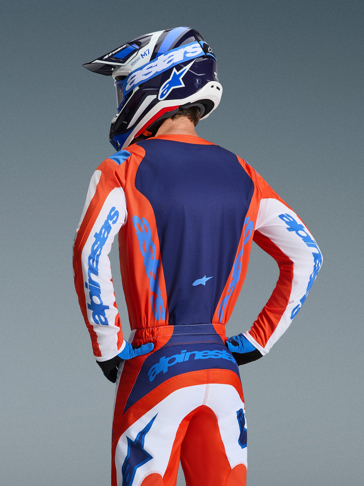Koszulka Alpinestars 2026 Fluid Grid, koszulka motocrossowa, pomarańczowa i niebieska (Ucla Blue), widok z tyłu motocyklisty w pasujących spodniach i kasku, z granatowym panelem tylnym i białymi rękawami z jasnoniebieskim brandingiem, sportowy krój do wyścigów terenowych
