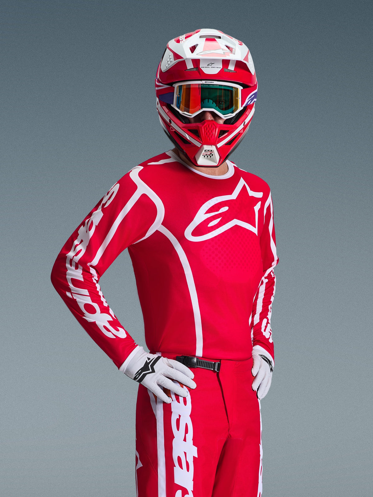 Motocyklista motocrossowy ubrany w zestaw Alpinestars 2026 Fluid Apex w kolorze czerwonym i białym, czerwona koszulka z białymi logotypami, pasujące spodnie, białe rękawice oraz czerwono-biały kask z opalizującymi goglami, kompletny strój do wyścigów terenowych