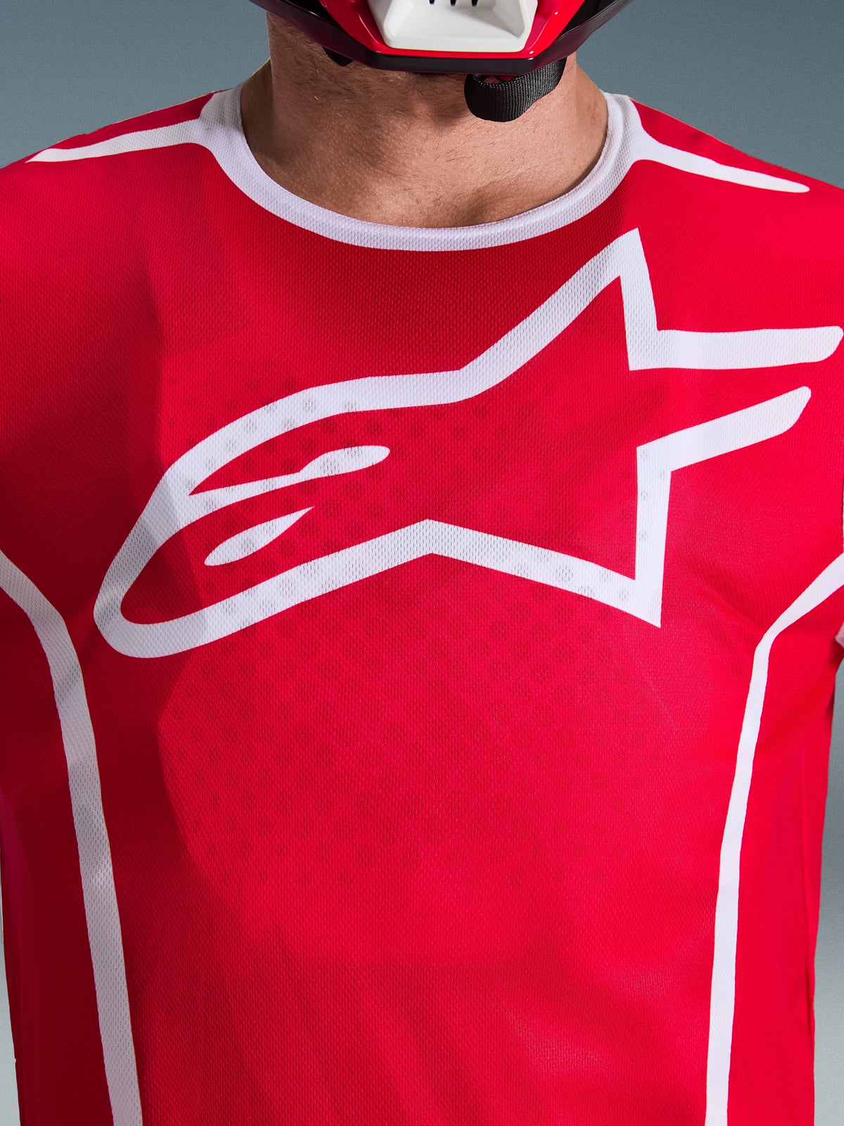 Zbliżenie na koszulkę Alpinestars 2026 Fluid Apex w kolorze czerwono-białym, z dużym białym logo Astars na klatce piersiowej, oddychającą konstrukcją z siatki poliestrowej i białym wykończeniem kołnierzyka, odzież motocrossowa
