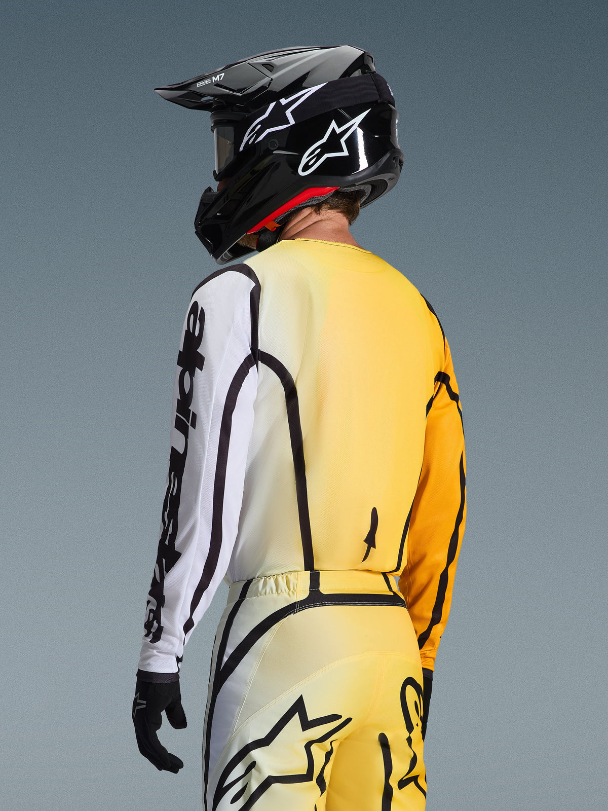 Widok z tyłu pod kątem trzech czwartych na zawodnika motocrossowego ubranego w koszulkę 2026 Fluid Apex w kolorze Yellow Black, z gradientem od żółtego do białego, wyraźnym czarnym brandingiem Alpinestars na rękawie, w zestawie z pasującymi spodniami i czarnym kaskiem motocrossowym.