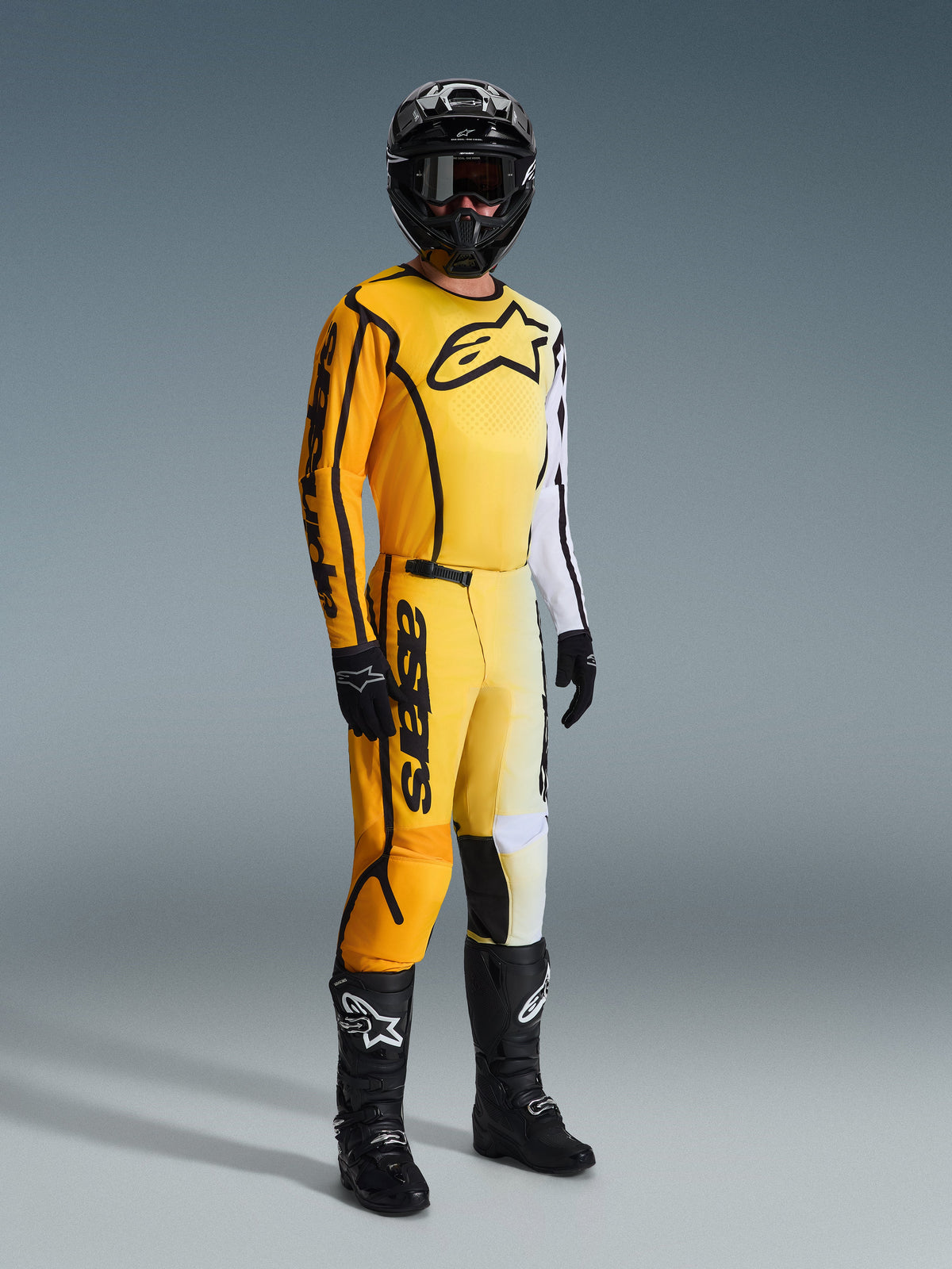 Motocyklista motocrossowy w koszulce Alpinestars 2026 Fluid Apex w kolorze żółto-czarnym, z gradientem od żółtego do białego i dużym czarnym logo na klatce piersiowej, dopasowane spodnie, czarny kask i czarne buty, kompletny strój wyścigowy do motocrossu off-road