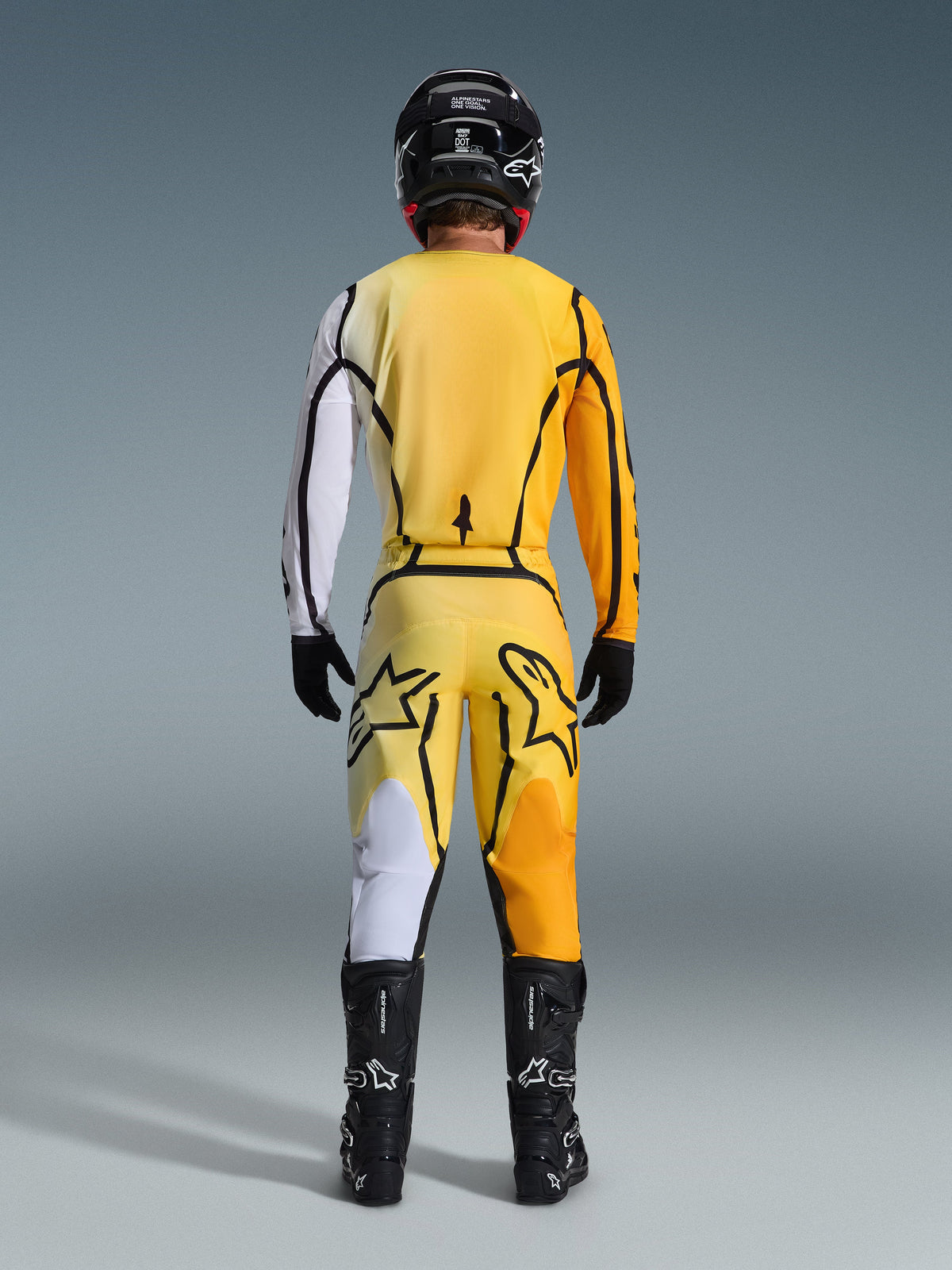 Motocross rider from behind wearing the Alpinestars 2026 Fluid Apex Gear Set, yellow jersey with a white sleeve and black accents, matching yellow pants featuring large black Alpinestars logos on the rear, black helmet, black gloves, and black boots, coordinated outfit for off-road racing