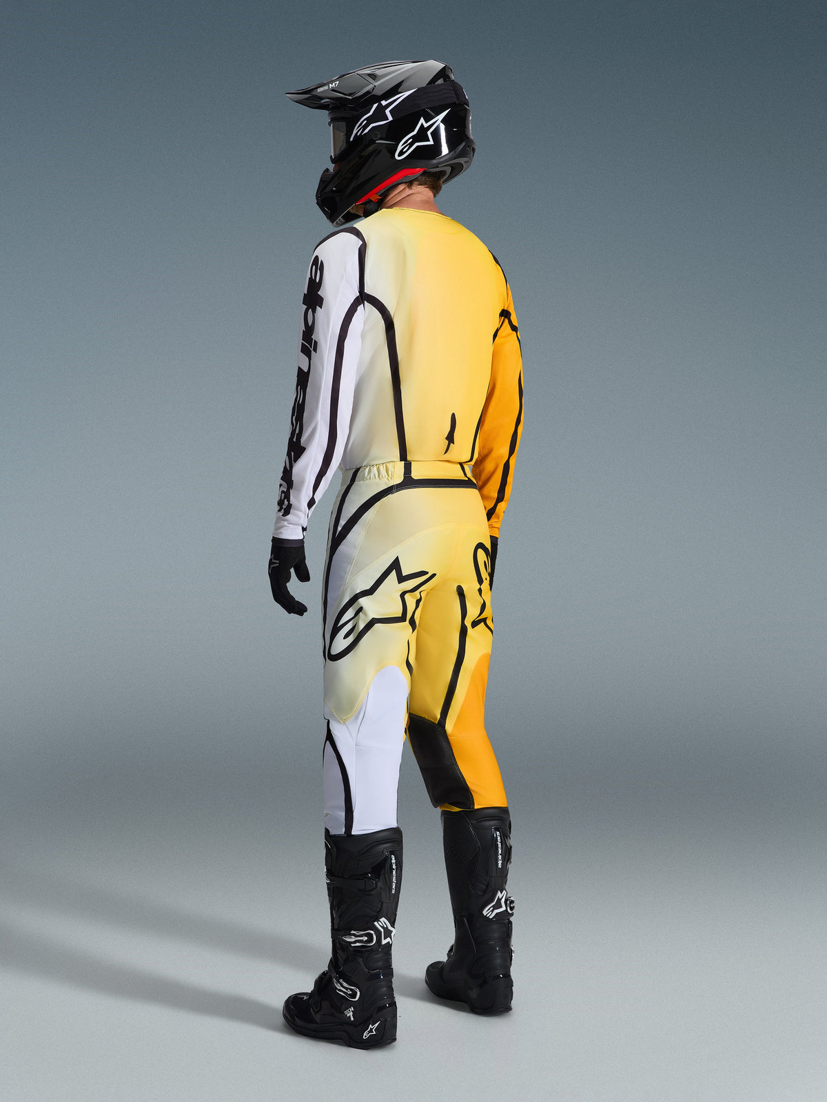 Motocross rider seen from behind wearing the Alpinestars 2026 Fluid Apex Gear Set in Yellow and Black, featuring an asymmetrical color-block jersey with white and orange-yellow panels, matching motocross pants, black helmet with white logo, black boots, and black gloves.