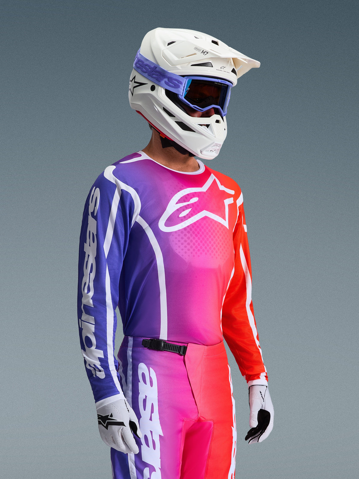 Koszulka Alpinestars 2026 Fluid Apex, koszulka motocrossowa, żywy gradient fioletu, różu i pomarańczu, noszona z pasującymi spodniami, biały kask z niebieskimi goglami i białe rękawice, sportowy design dla wydajności off-road i mobilności