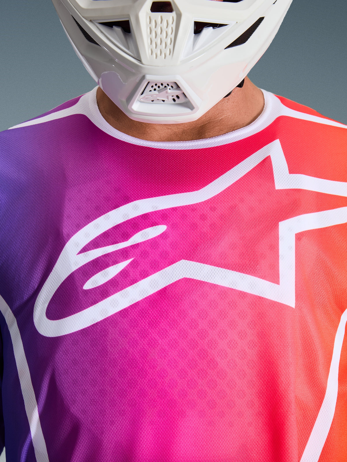 Alpinestars 2026 Fluid Apex Jersey, Motocross Jersey, Multicolor Purple Pink and Orange Gradient, featuring a large white Astars logo on the chest, close-up view worn with a white motocross helmet