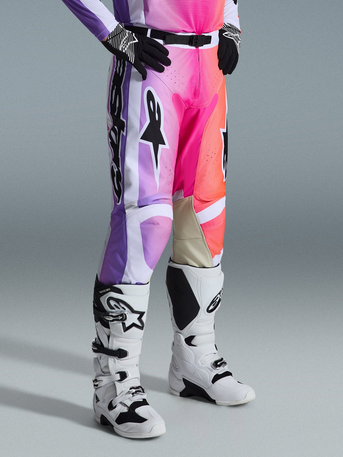 Alpinestars 2026 Racer Air Portl Pants, spodnie motocrossowe, wielokolorowa grafika z fioletowymi, różowymi i pomarańczowymi gradientami, noszone z biało-czarnymi butami motocrossowymi i czarnymi rękawicami, sportowy krój race fit i jasnobrązowe skórzane panele po wewnętrznej stronie kolan.