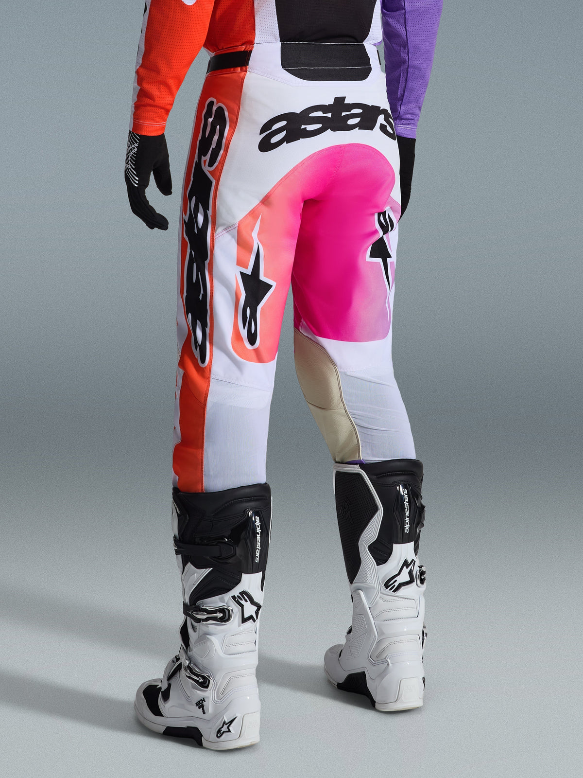 Alpinestars 2026 Racer Air Portl Pants, spodnie motocrossowe, wielokolorowe z białymi, jaskraworóżowymi i pomarańczowymi elementami, widok z tyłu w biało-czarnych butach Alpinestars, wyraźny branding i wytrzymała konstrukcja wyścigowa