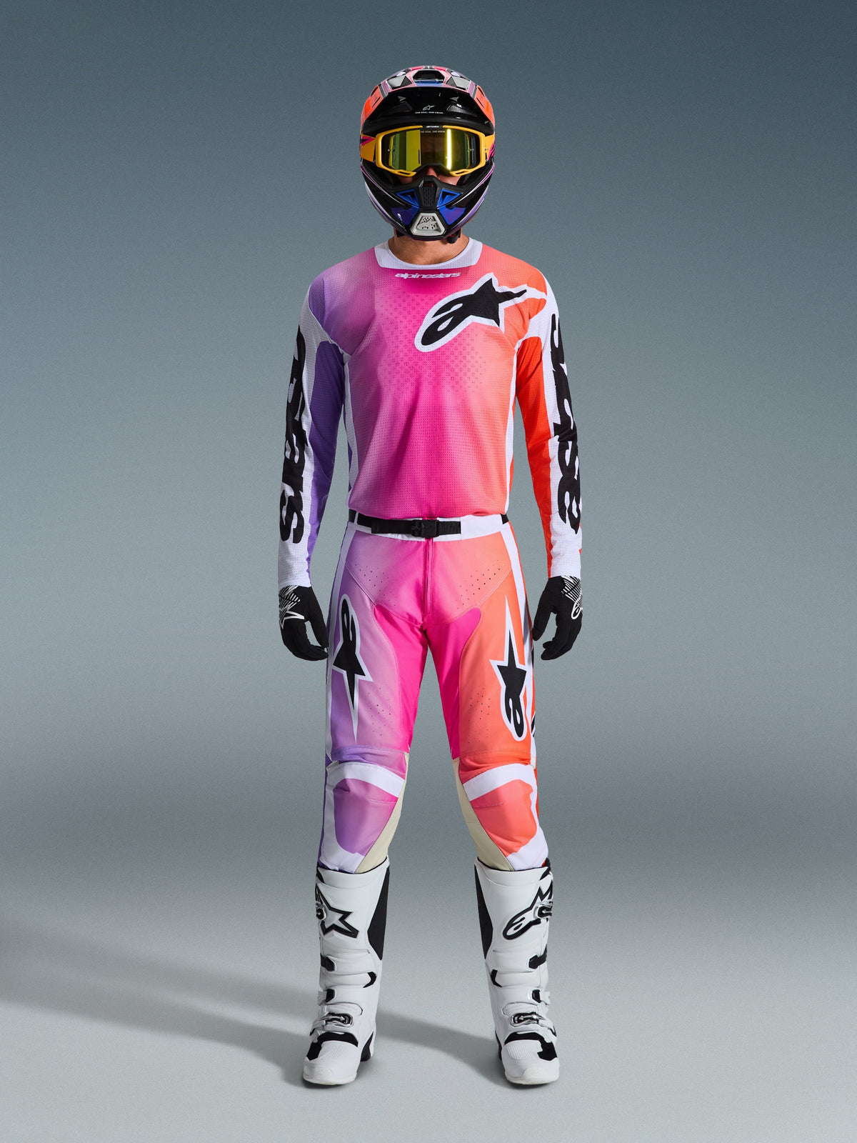 Zawodnik motocrossowy w pełnym stroju wyścigowym Alpinestars 2026 Racer Air Portl z różowo-fioletowo-pomarańczowym gradientem na spodniach i koszulce, w dopasowanym kasku, żółtych goglach, białych butach i czarnych rękawicach, zapewniających spójny wygląd off-road.