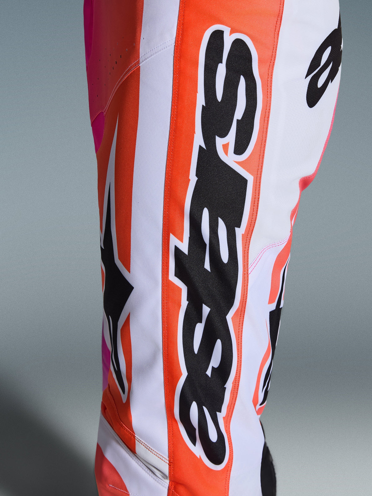 Alpinestars Racer Air Portl Pants 2026, spodnie motocrossowe, pomarańczowe, białe i różowe, zbliżenie na duże czarne logo Alpinestars i gwiazdę, perforowany materiał zapewniający wentylację, wytrzymała odzież do jazdy terenowej