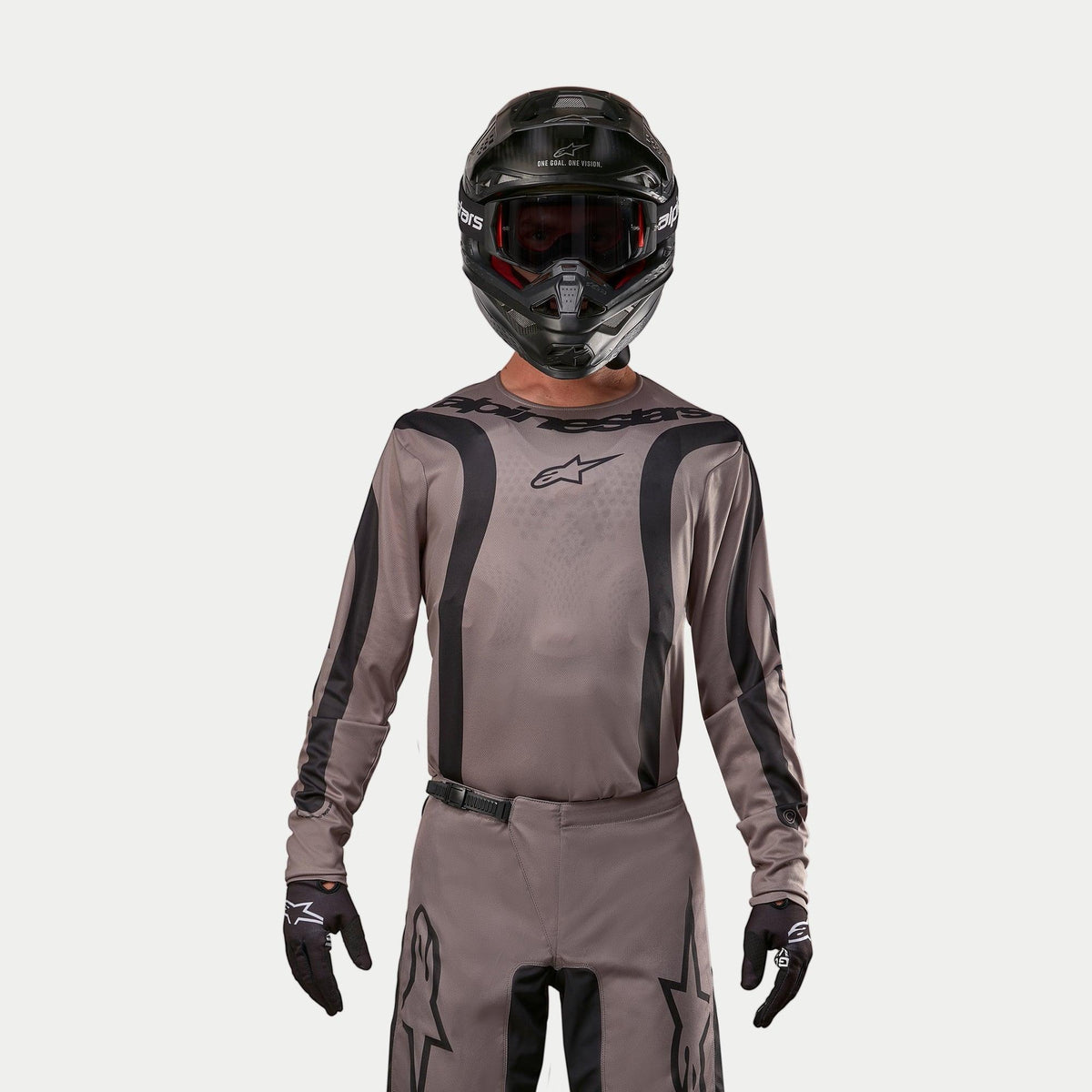 Motocyklista motocrossowy w koszulce Alpinestars 2024 Fluid Lurv w kolorze Mud Black, wykonanej z ziemistej szarobrązowej tkaniny z czarnymi graficznymi pasami i logo gwiazdy na piersi, w połączeniu z czarnym kaskiem, czerwonymi goglami i czarnymi rękawicami dla spójnego wyścigowego wyglądu off-road
