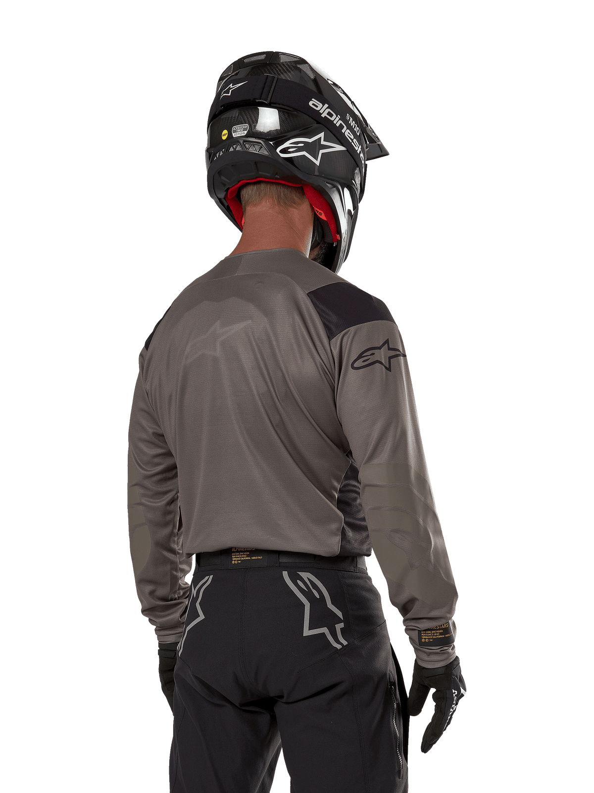 Motocyklista motocrossowy widoczny od tyłu ubrany w koszulkę Alpinestars Techdura w kolorze Falcon Brown, czarne spodnie terenowe i czarny kask, nowoczesny design graficzny, wydłużony tył do jazdy enduro