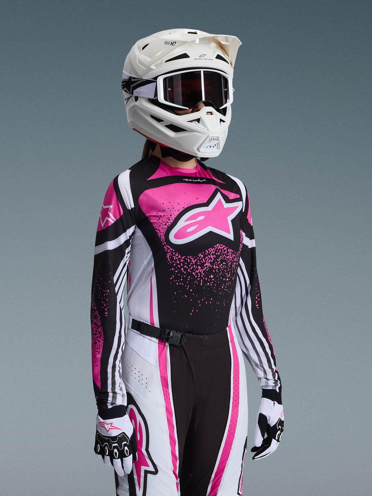 Zawodniczka motocrossowa w zestawie Alpinestars 2026 Stella Techstar Nomur w kolorze różowym, czarnym i białym; koszulka z dużym różowym logo gwiazdy, spodnie do kompletu z klamrą magnetyczną, biały kask z goglami oraz biało-czarne rękawice.