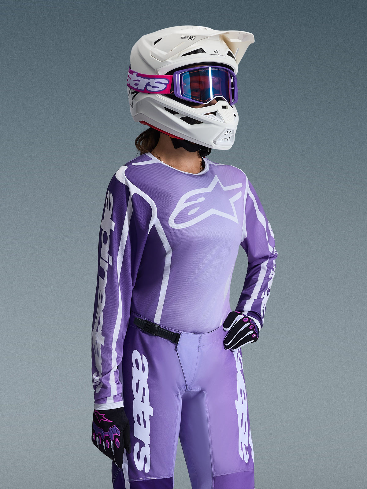 Zawodniczka motocrossowa w zestawie Alpinestars 2026 Stella Fluid Apex, fioletowo-biała bluza z logo Alpinestars, pasujące fioletowe spodnie, biały kask z fioletowymi goglami oraz czarno-fioletowe rękawice, profesjonalny strój off-road