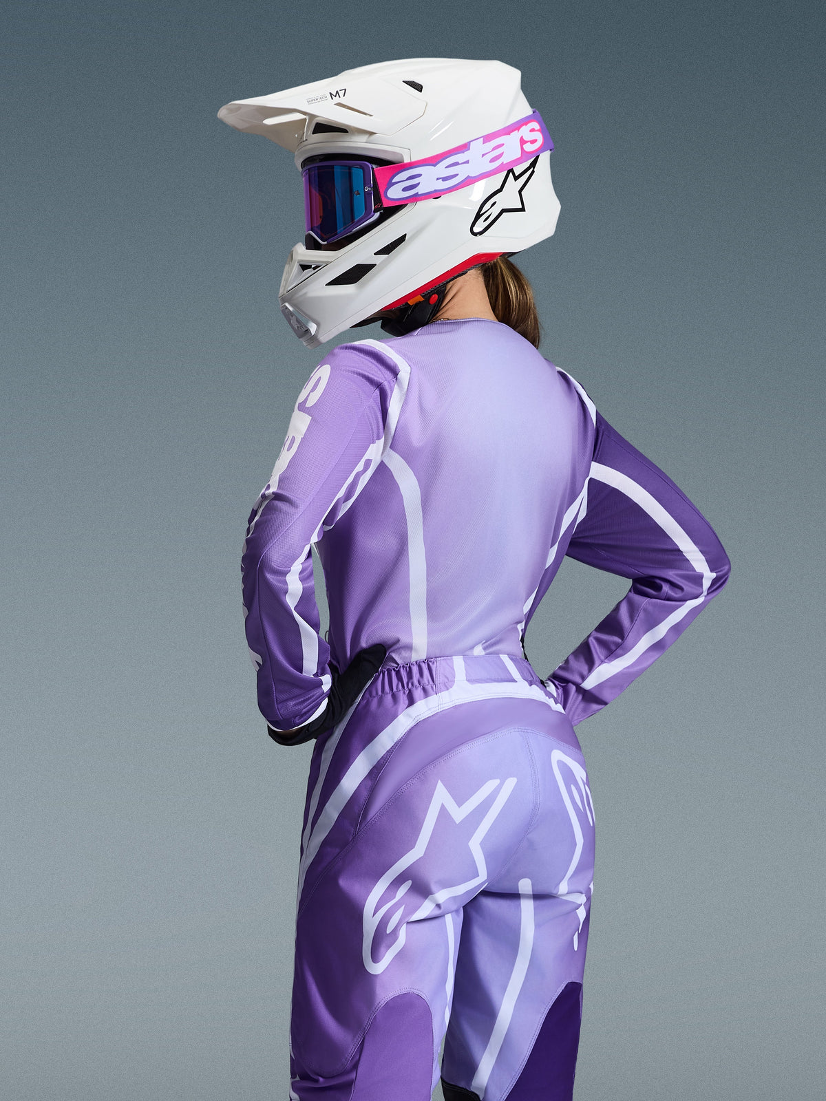 Zawodniczka motocrossowa w zestawie Alpinestars 2026 Stella Fluid Apex, fioletowa i lawendowa koszulka oraz spodnie z białą grafiką, biały kask z różowo-fioletowymi goglami, dedykowany damski krój, dopasowany strój do wyścigów off-road