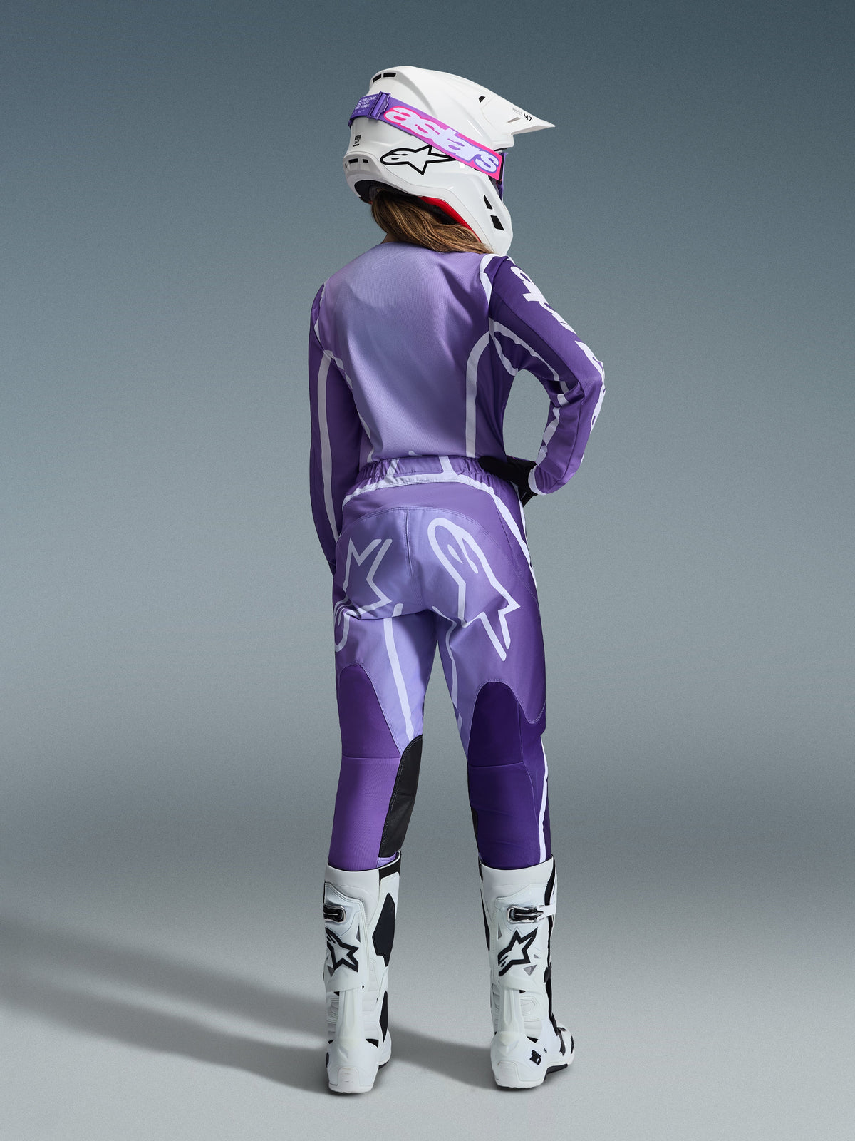Alpinestars 2026 Stella Fluid Apex Gear Set, damska odzież motocrossowa, fioletowy i lawendowy z białymi akcentami, motocyklistka widziana od tyłu w dopasowanej koszulce i spodniach, biały kask z różowymi goglami i białe buty, skoordynowany sportowy strój wyścigowy