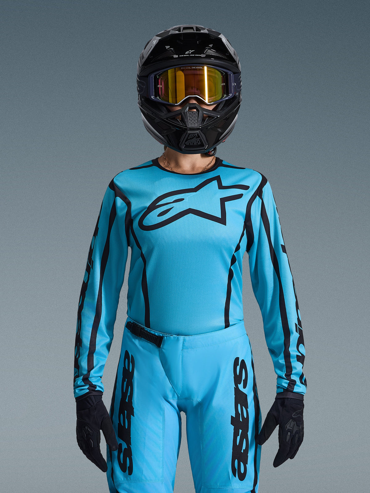 Zawodniczka motocrossowa w damskim zestawie Alpinestars 2026 Stella Fluid Apex, jasnoniebieska koszulka z czarnymi konturami i logo, pasujące spodnie, czarny kask z opalizującymi złotymi goglami i czarne rękawice, dopasowany damski strój wyścigowy do motocrossu