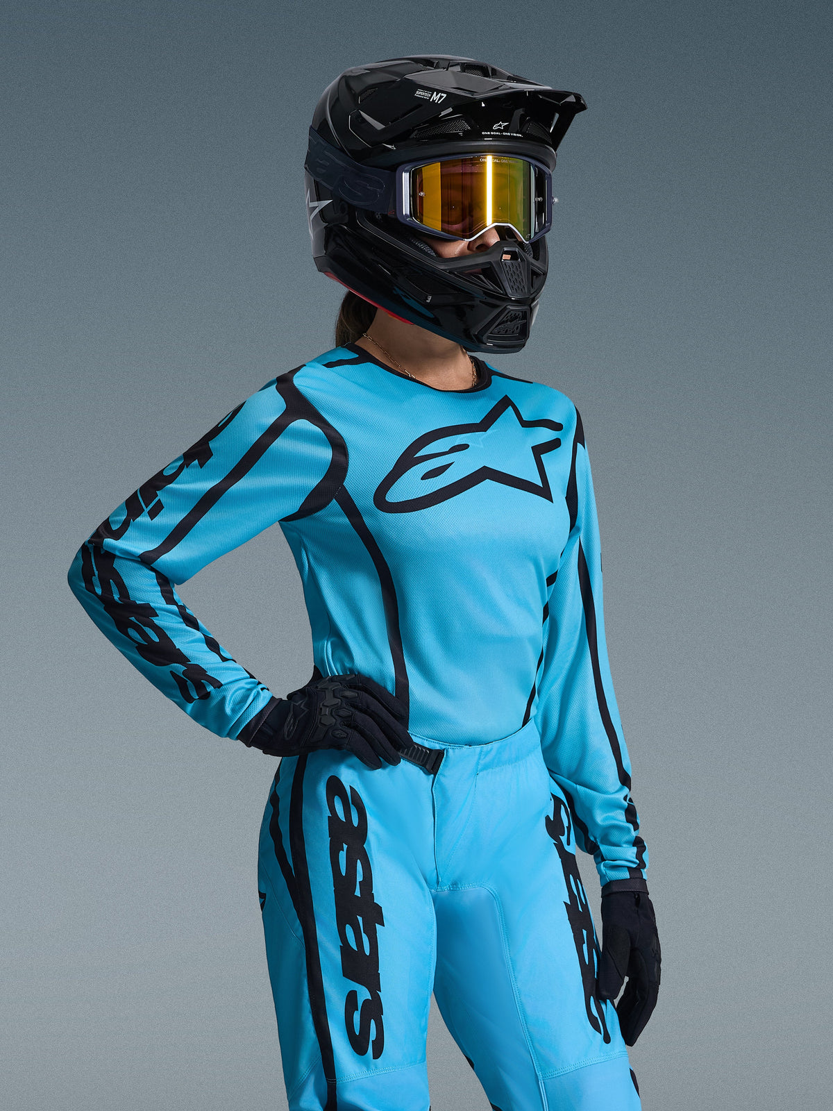 Damski zestaw Alpinestars Stella Fluid Apex 2026, jasnoniebieski i czarny, koszulka i spodnie motocrossowe z czarnym logowaniem, czarny kask, złote lustrzane gogle i czarne rękawice, dedykowany krój damski do wyścigów off-road