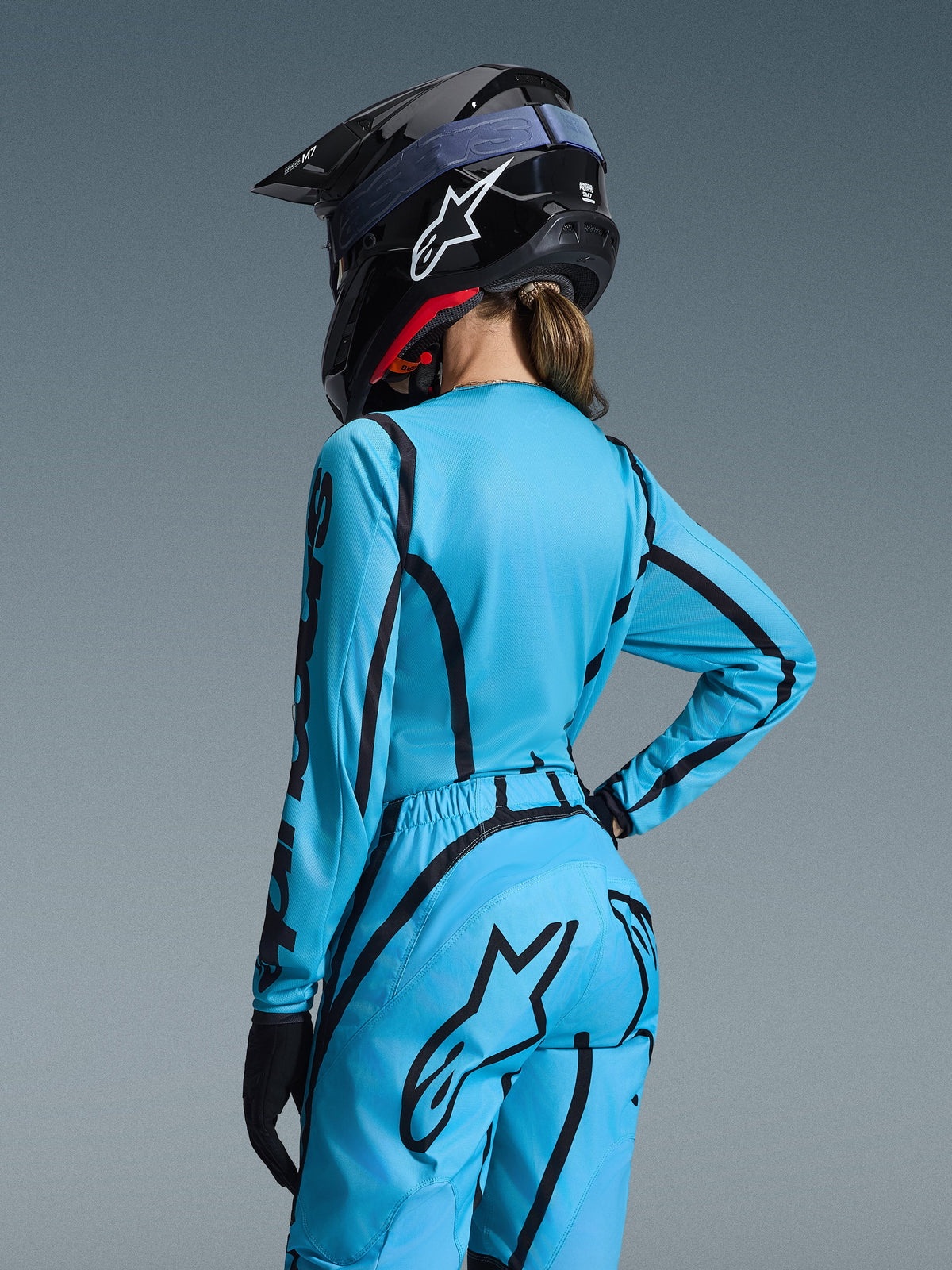Alpinestars Stella Fluid Apex 2026 Gear Set, jasnoniebieski i czarny, widok z tyłu na motocyklistkę w koszulce motocrossowej i dopasowanych spodniach, czarny kask Alpinestars z białym logo, techniczna odzież wyścigowa off-road dla kobiet