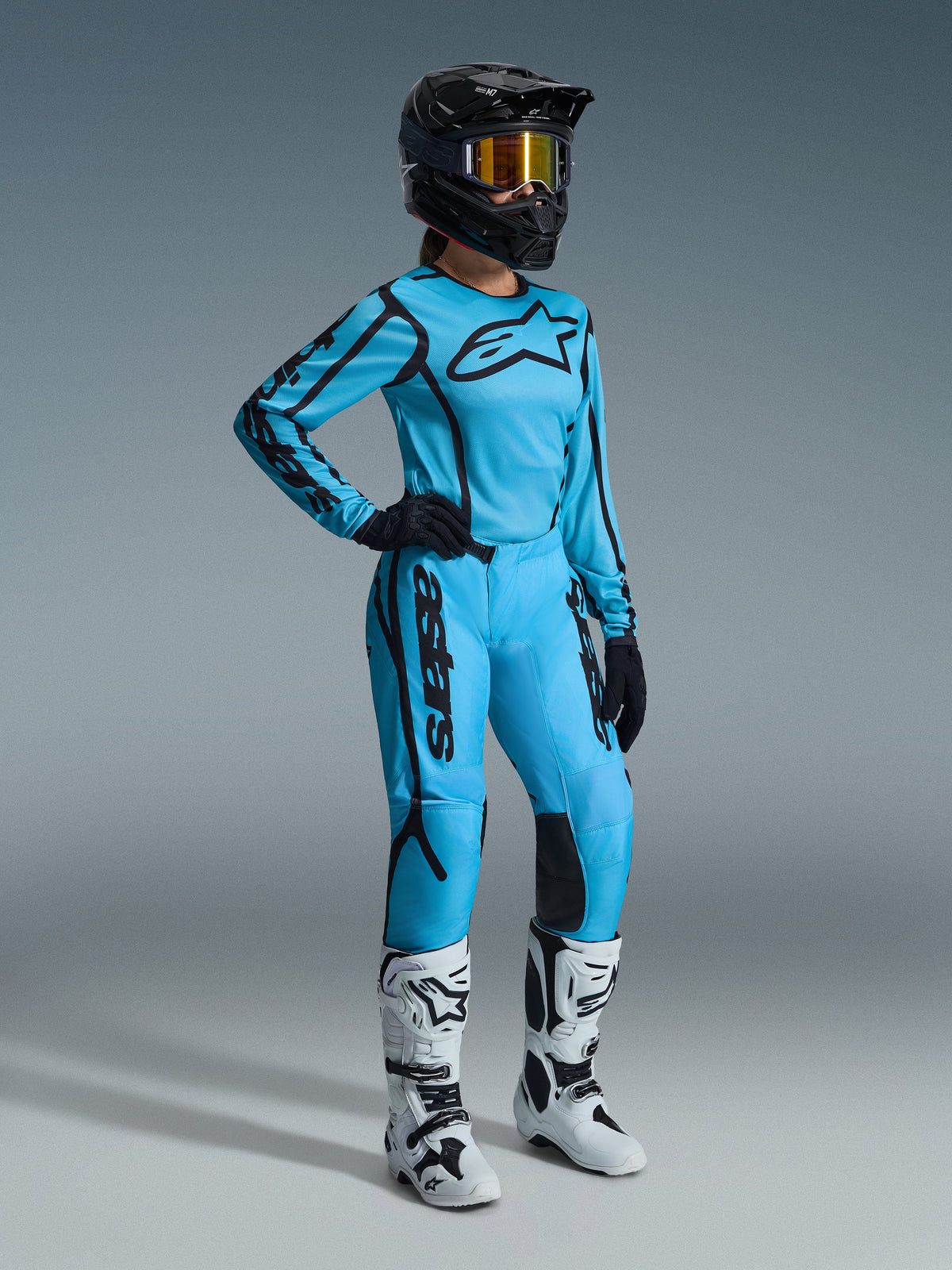 Damski zestaw Alpinestars Stella Fluid Apex 2026 w kolorze Tropical Black, jasnoniebieska koszulka i pasujące spodnie z czarnymi akcentami, na zawodniczce w czarnym kasku, opalizujących goglach i białych butach, profesjonalny strój motocrossowy.