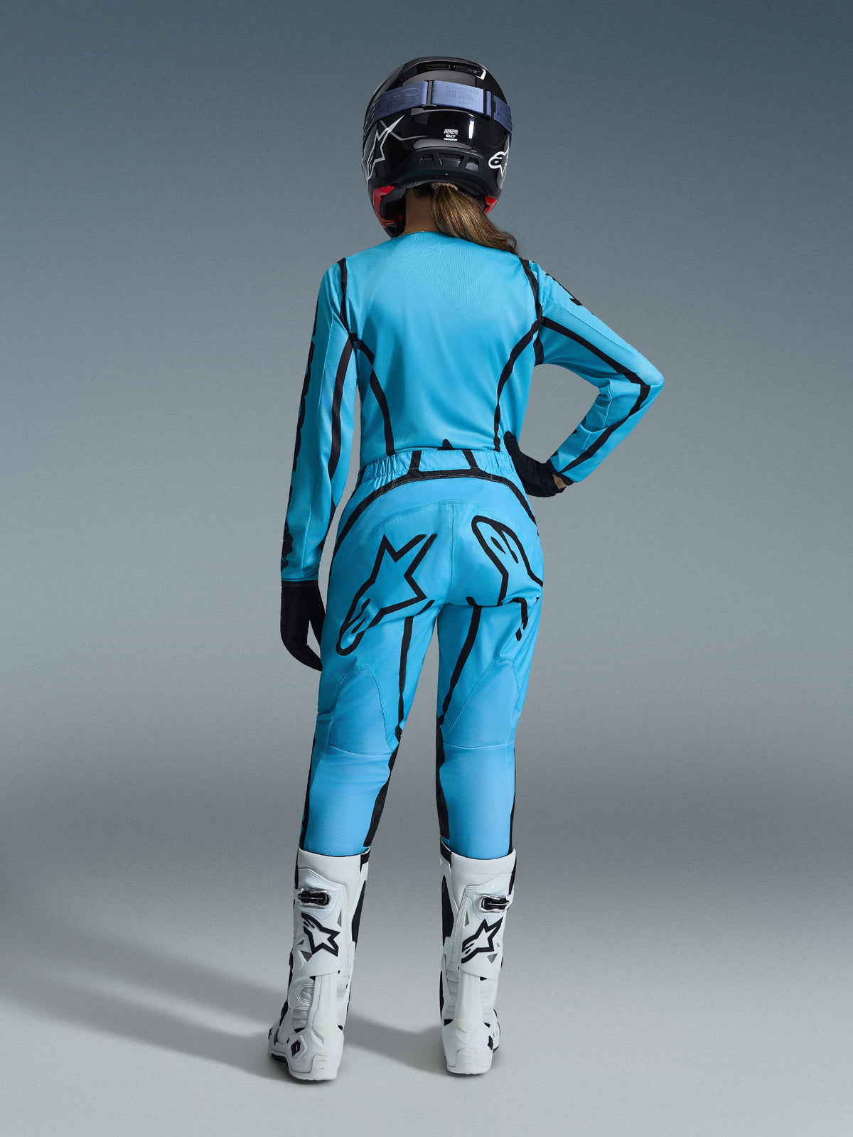 Zawodniczka motocrossowa widoczna od tyłu, ubrana w jaskrawoniebieski zestaw Alpinestars 2026 Woman Stella Fluid Apex z czarnymi akcentami graficznymi, z dużym czarnym logo gwiazdy na spodniach, biało-czarnymi butami i czarnym kaskiem.