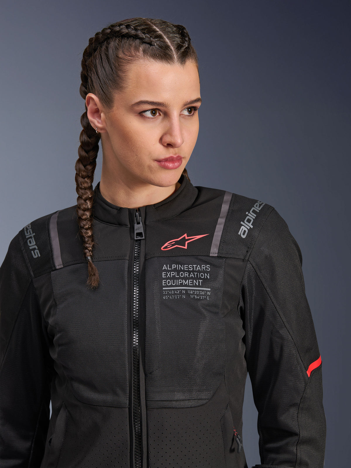 Alpinestars Stella ST-2 Air Jacket, damska kurtka motocyklowa w kolorze czarnym, z panelami z siatki zapewniającymi przepływ powietrza, brandingiem „Alpinestars Exploration Equipment” ze współrzędnymi i czerwonym logo na klatce piersiowej, prezentowana przez modelkę z warkoczami.