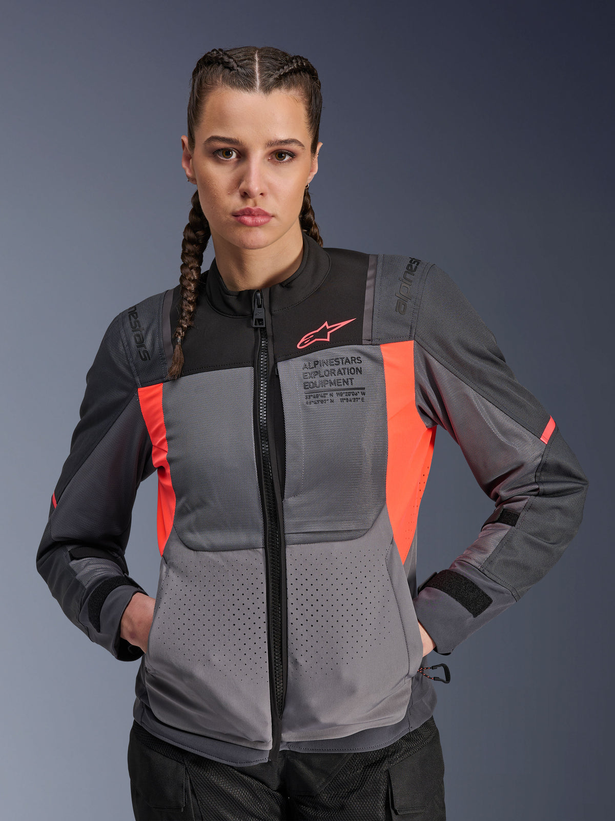 Kurtka Alpinestars Stella ST-2 Air, damska kurtka motocyklowa, ciemnoszara z koralowymi elementami fluo, noszona przez modelkę z warkoczami, wyposażona w techniczne panele z siatki zapewniające przepływ powietrza, laserowe perforacje oraz branding „Alpinestars Exploration Equipment” na klatce piersiowej, letnia odzież turystyczna