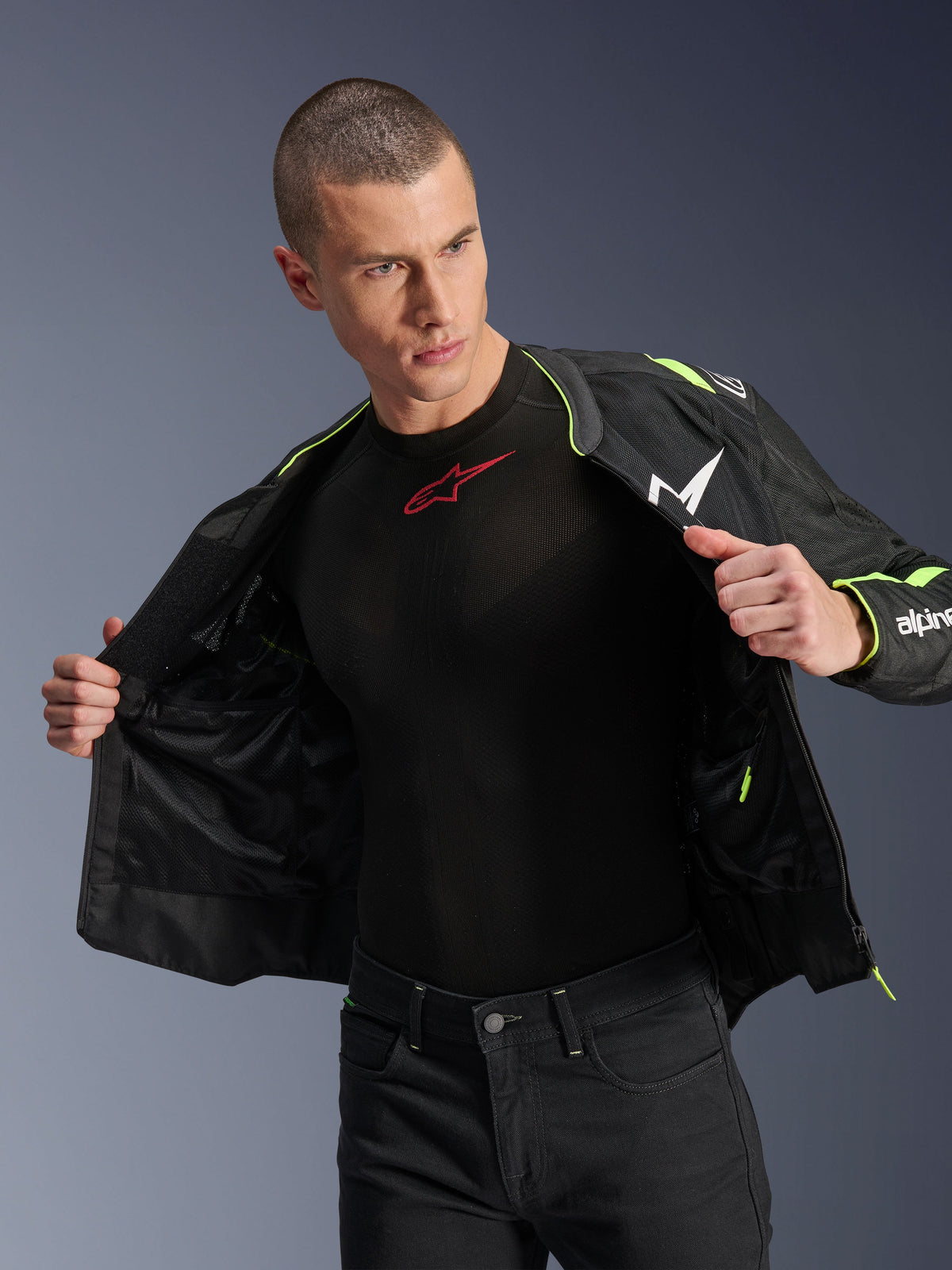 Męski model ubrany w kurtkę Alpinestars T-Sps Air V2 w kolorze czarnym i żółtym fluo, rozpiętą, aby wyeksponować wewnętrzną konstrukcję z siatki i kieszenie, w zestawieniu z czarną warstwą bazową Alpinestars i czarnymi spodniami, miejski styl odzieży motocyklowej