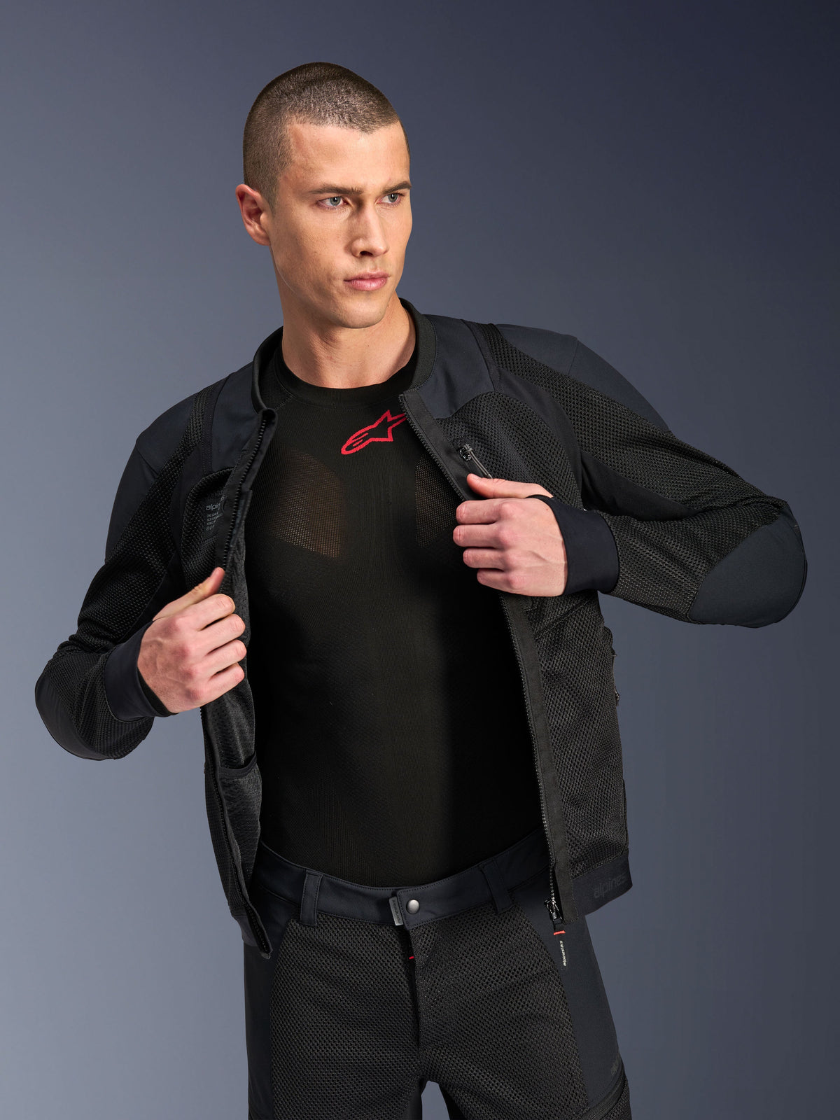 Kurtka Alpinestars Troop-Air Pro, czarna, techniczna kurtka ochronna premium o siateczkowej konstrukcji, noszona rozpięta na czarnej warstwie bazowej z czerwonym logo, zestawiona z pasującymi siateczkowymi spodniami motocyklowymi, współczesny miejski styl jazdy