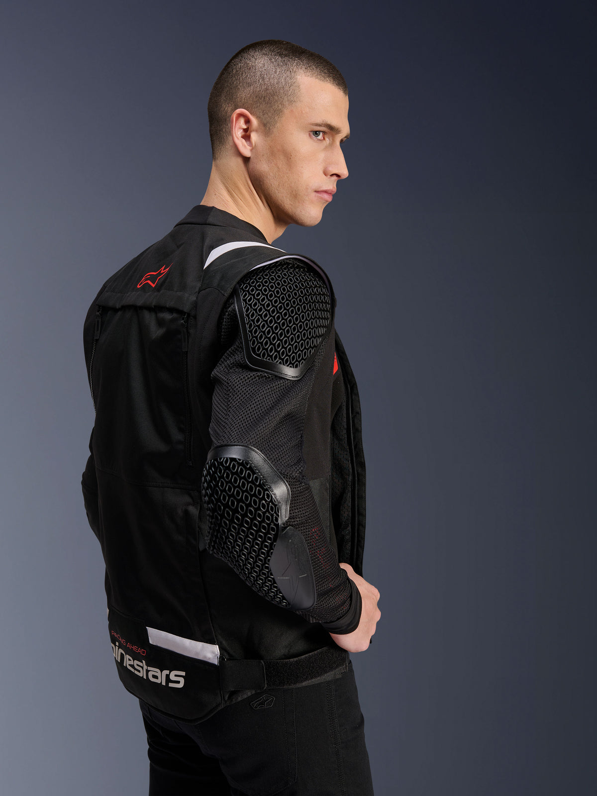 Alpinestars T-Stunt Air Jacket, kurtka motocyklowa, czarna, profil boczny modela w kurtce o sportowym kroju z siatkowymi rękawami, widoczne ochraniacze o strukturze plastra miodu na ramieniu i łokciu, czerwone logo na plecach, białe logo w pasie