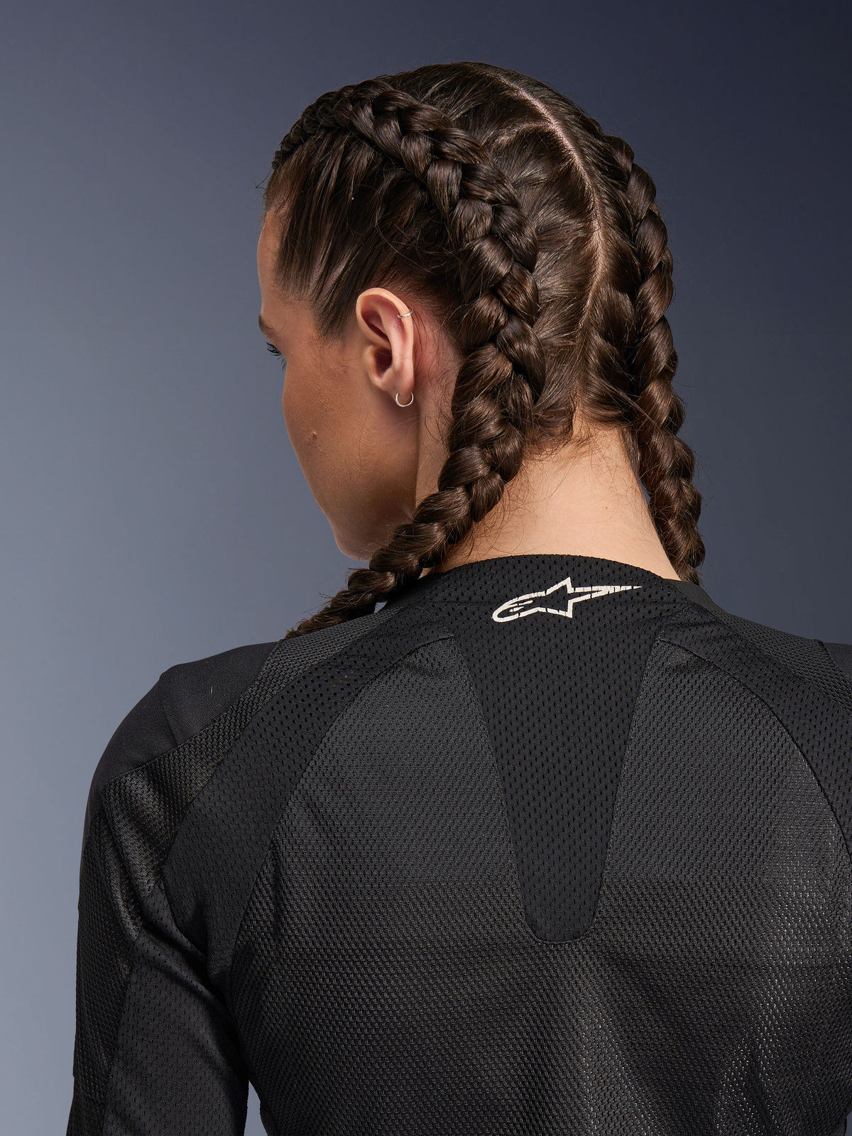Damska kurtka Alpinestars Stella Troop-Air, czarna, szczegółowy widok z tyłu na górną część pleców i kark, kobieta z włosami zaplecionymi w warkocze francuskie, białe logo Astars na kołnierzu, lekka techniczna siatka 2D zapewniająca wentylację