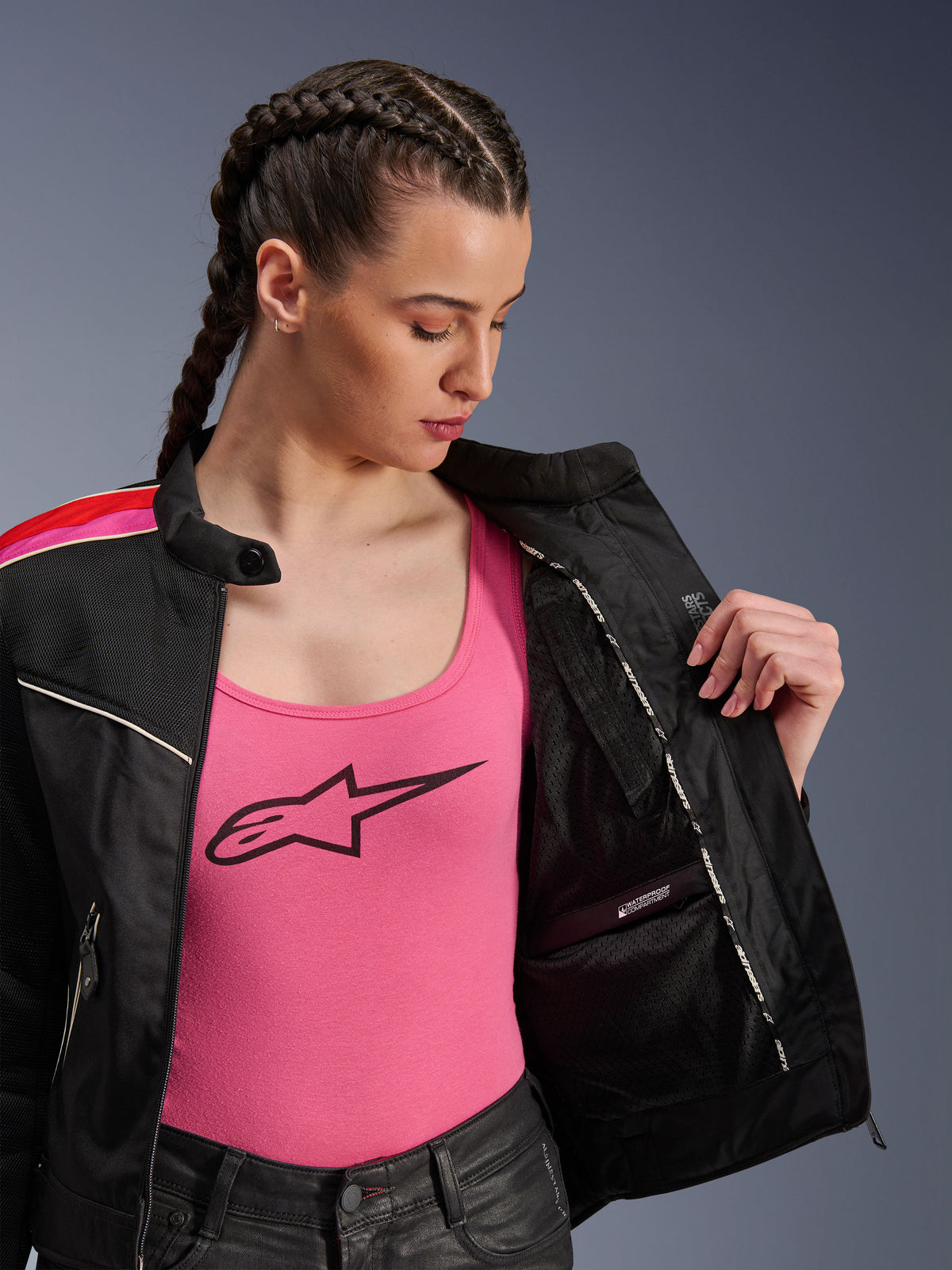 Motocyklistka ubrana w kurtkę Alpinestars Stella T-Dyno Air w kolorze Black Bright Red Fuchsia, częściowo rozpiętą, aby odsłonić wewnętrzną wodoodporną przegrodę, założoną na różowy top z logo, co podkreśla oddychające panele z siatki i dopasowany motocyklowy krój.