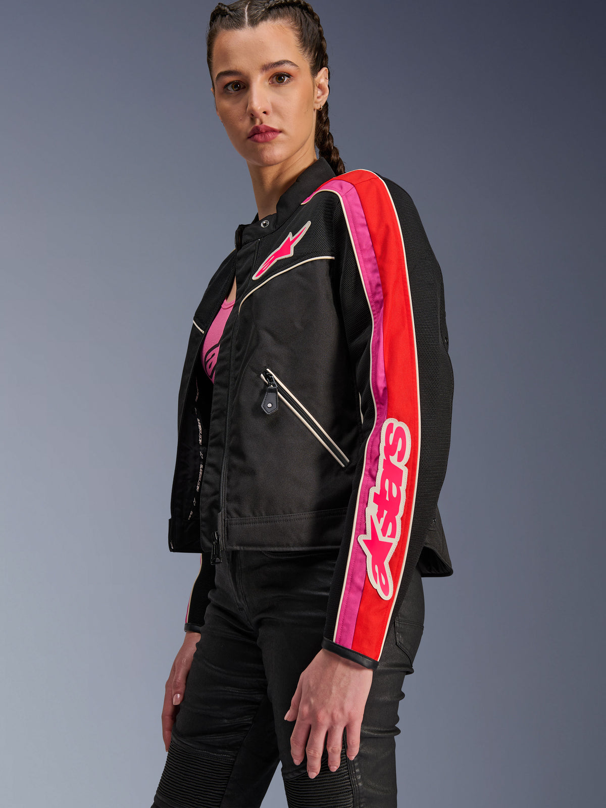 Alpinestars Stella T-Dyno Air Jacket, damska kurtka motocyklowa, czarna z jaskrawoczerwonymi i fuksjowymi akcentami, noszona z czarnymi spodniami, z różowym logo Astars na piersi i rękawie, taliowany motocyklowy krój z panelami z siatki zapewniającymi wentylację