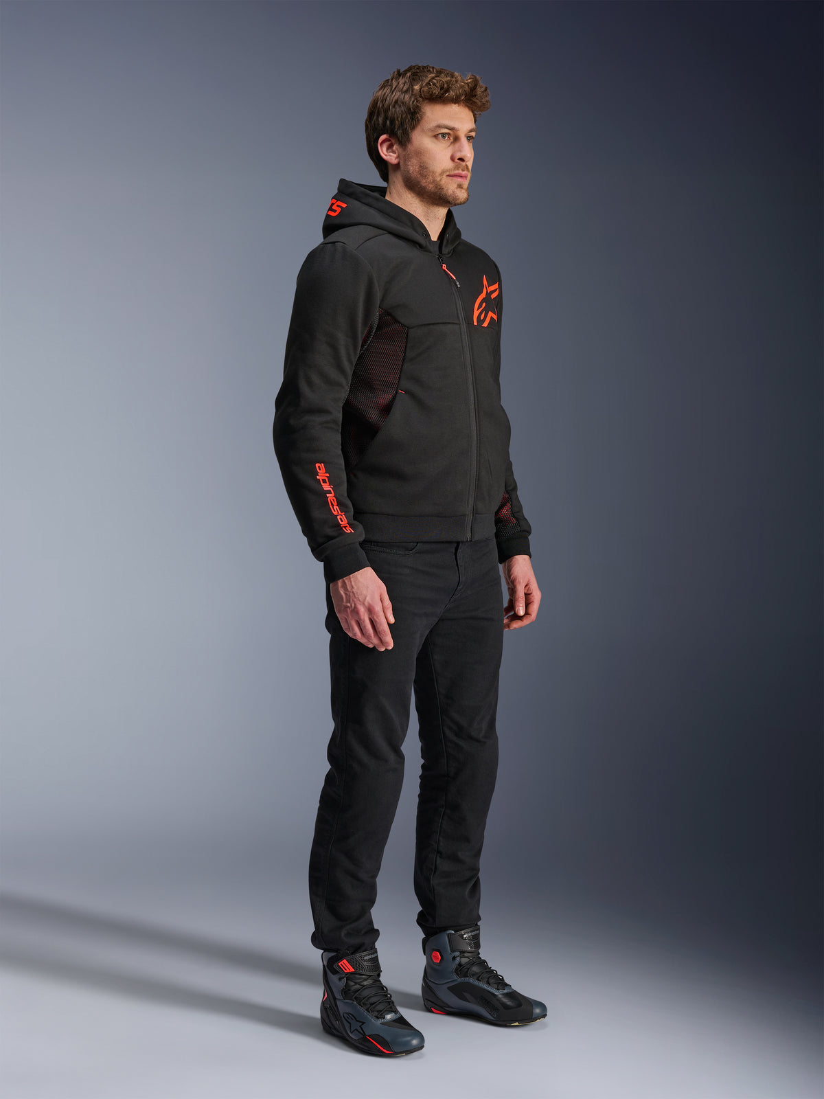 Alpinestars Chrome Air Sport Hoodie, Black Red Fluo, miejska bluza motocyklowa z technicznego polaru, noszona z czarnymi spodniami i butami motocyklowymi, wyposażona w siatkowe panele wentylacyjne i czerwone fluorescencyjne logo na klatce piersiowej i rękawie.