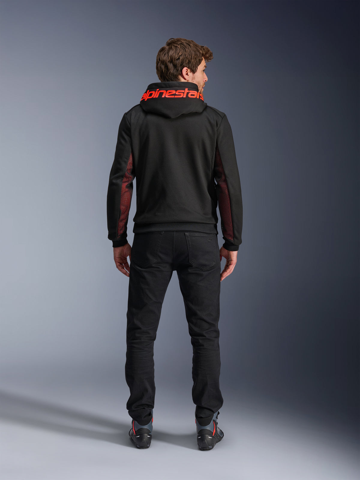 Alpinestars Chrome Air Sport Hoodie, czarno-czerwona (Black Red Fluo), widok z tyłu na mężczyznę w miejskiej kurtce motocyklowej, czerwony napis Alpinestars na kapturze, czerwone panele z siatki na rękawach, czarne spodnie