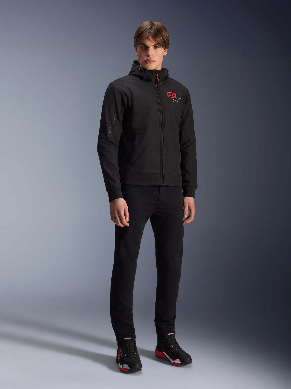 Alpinestars MM93 Radium Tech Hoodie, miejska kurtka motocyklowa, czarna z jaskrawoczerwonymi akcentami, prezentowana przez modela w czarnych spodniach i butach motocyklowych, z czerwonym logo „93” na piersi i brandingiem Marca Marqueza na rękawie, casualowa techniczna odzież uliczna do jazdy miejskiej