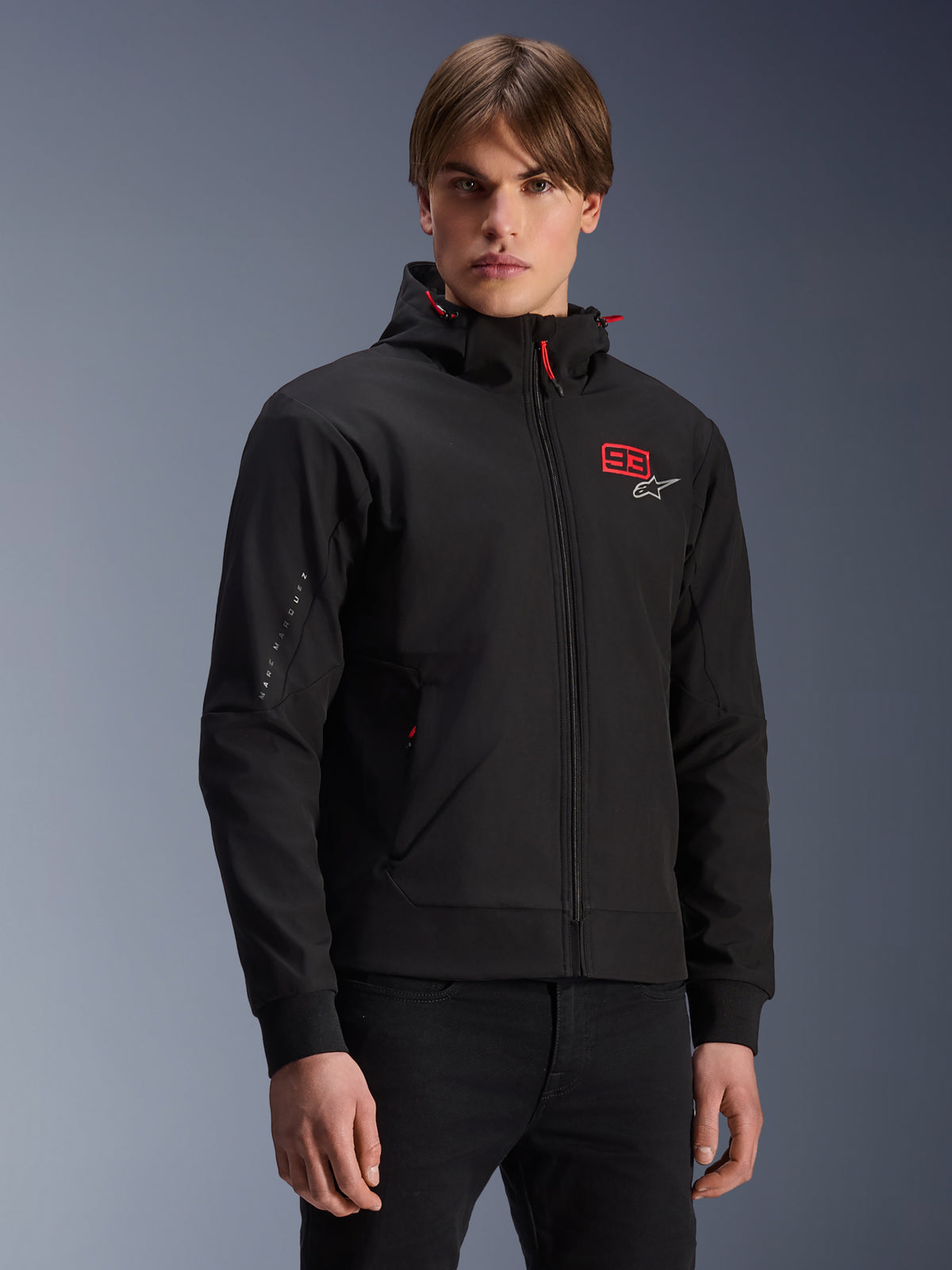 Bluza z kapturem MM93 Radium Tech, czarna miejska kurtka motocyklowa zapinana na zamek, czerwony numer 93 i białe logo Alpinestars na piersi, noszona przez modela w czarnych spodniach, odpinany kaptur z czerwonymi sznurkami, branding na rękawie, casualowy design inspirowany motorsportem