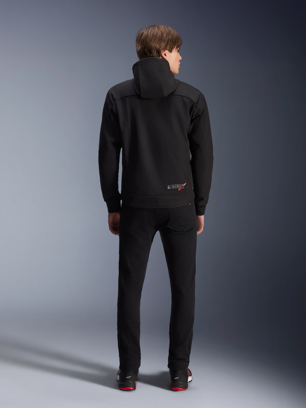 Alpinestars MM93 Radium Tech Hoodie, czarna, widok z tyłu na modelu, małe logo MM93 z czerwoną gwiazdą na dole pleców, zestawiona z czarnymi spodniami, miejska odzież motocyklowa
