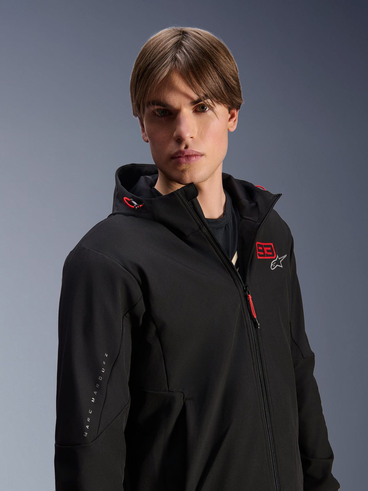 Męski model w bluzie Alpinestars MM93 Radium Tech Hoodie, czarna miejska kurtka motocyklowa z czerwonym logo „93” i białą gwiazdą na klatce piersiowej, brandingiem „Marc Marquez” na rękawie, techniczna wiatroodporna powłoka do jazdy miejskiej.
